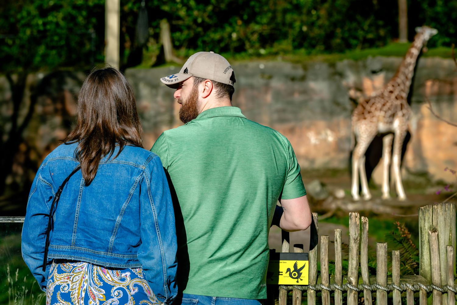 Two people in blue and green attire stand together against a rustic outdoor backdrop.