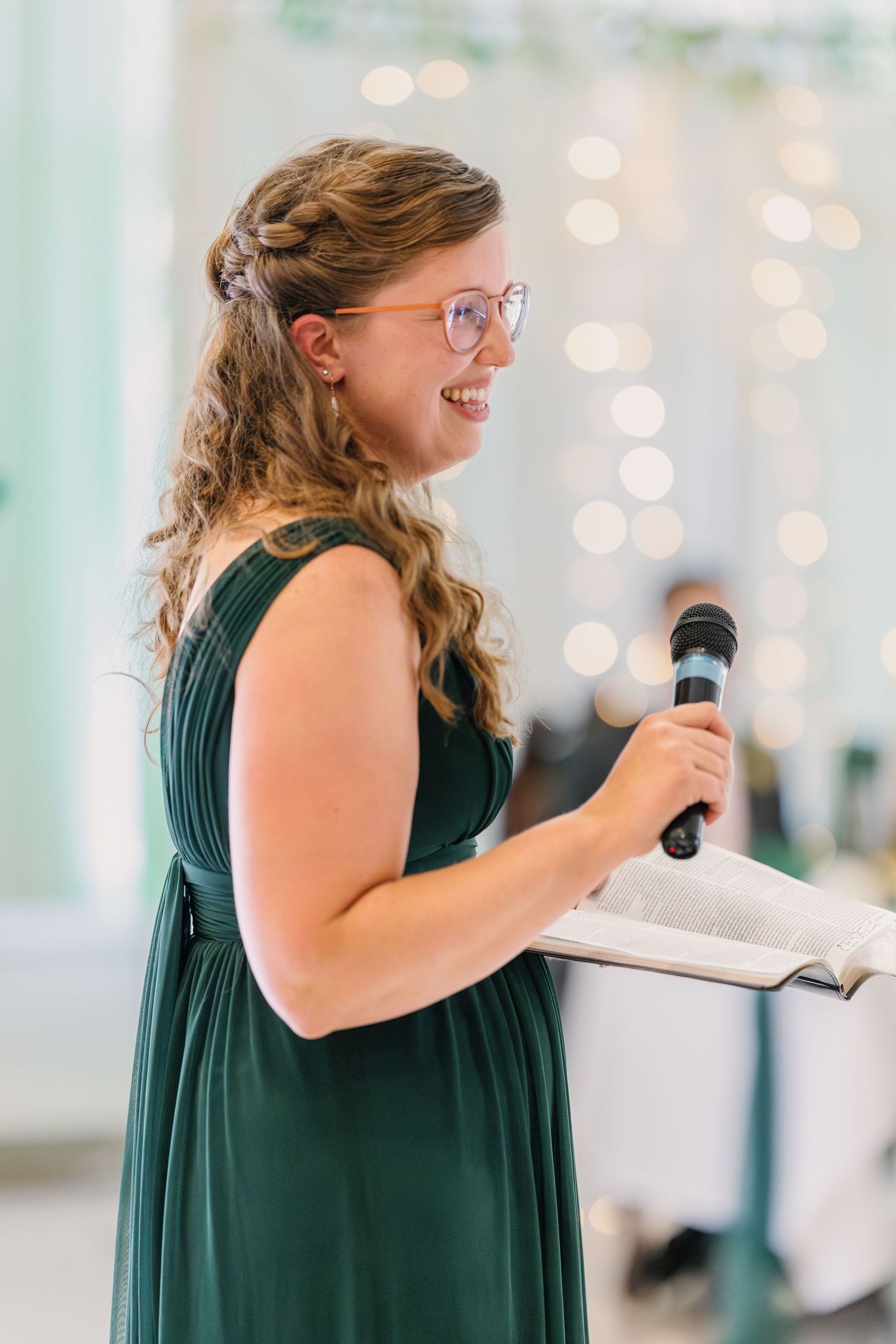 A bridesmaid in a dark green dress delivers a speech while holding a microphone and book at the wedding reception.