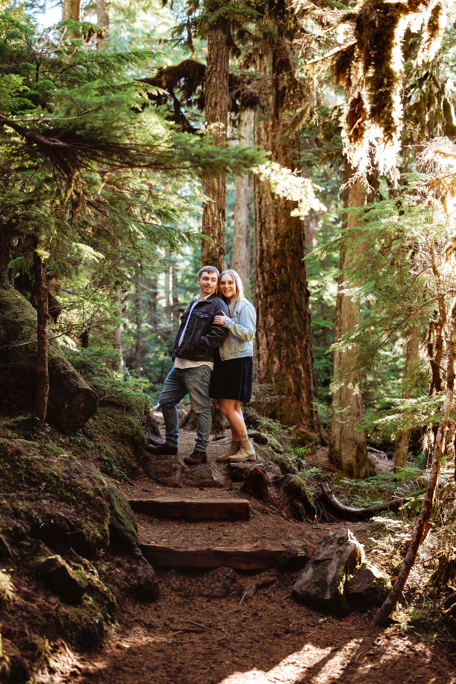 A couple walking along a shaded forest trail surrounded by tall evergreen trees.