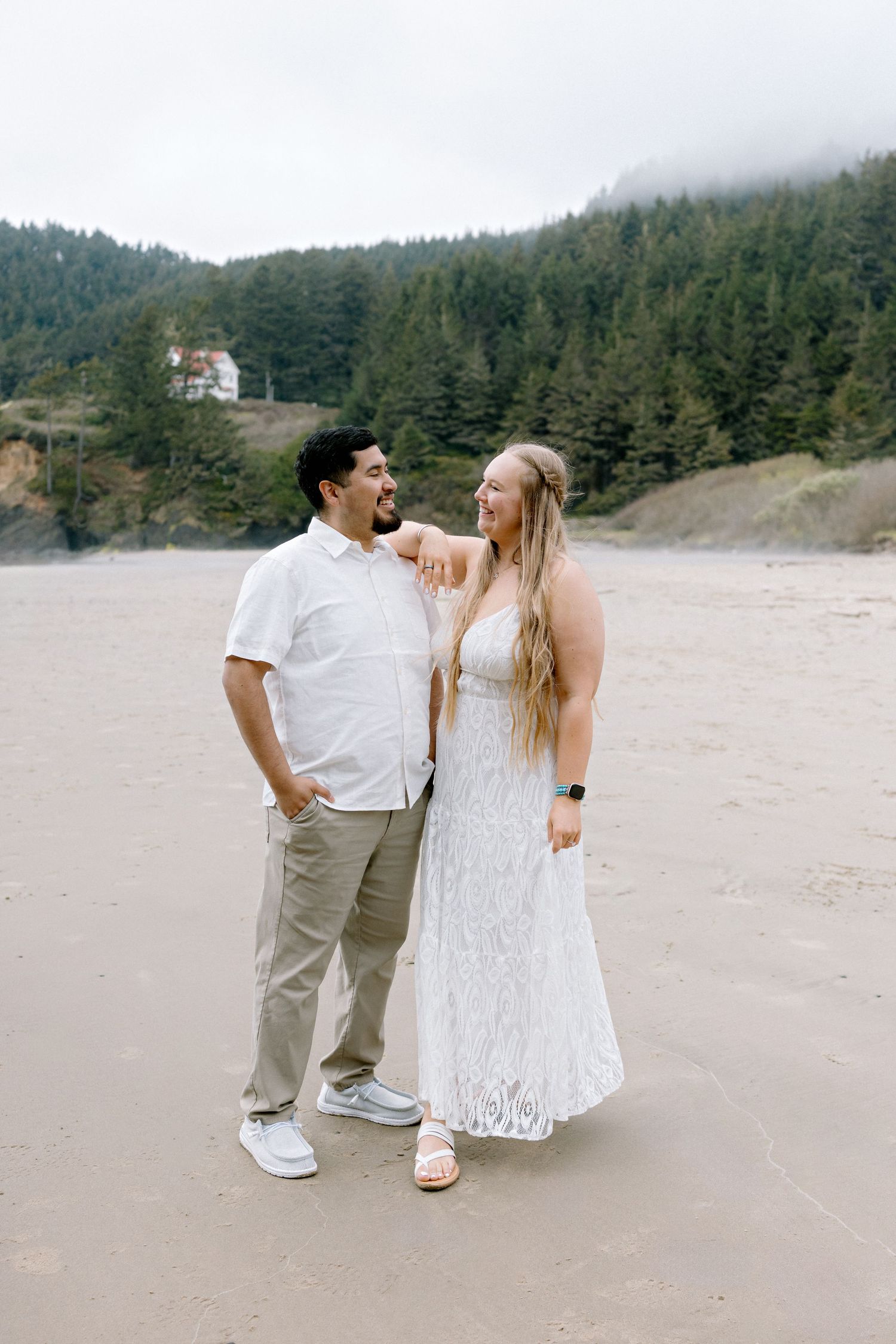 Couple in white outfits smiling on a sandy beach with misty forest and hills in the background.