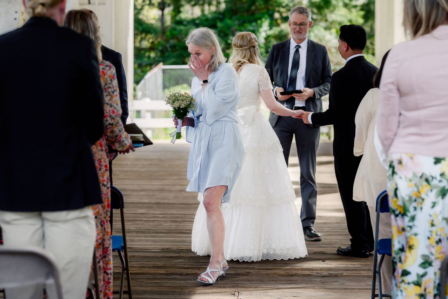 Emotional mother wipes tears as bride in white dress stands at outdoor wedding ceremony altar.