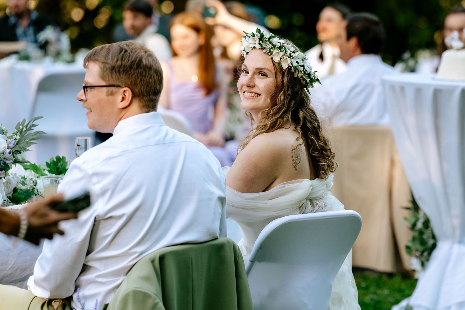 Bride in floral crown smiles warmly at outdoor wedding reception seated beside her groom at dusk.