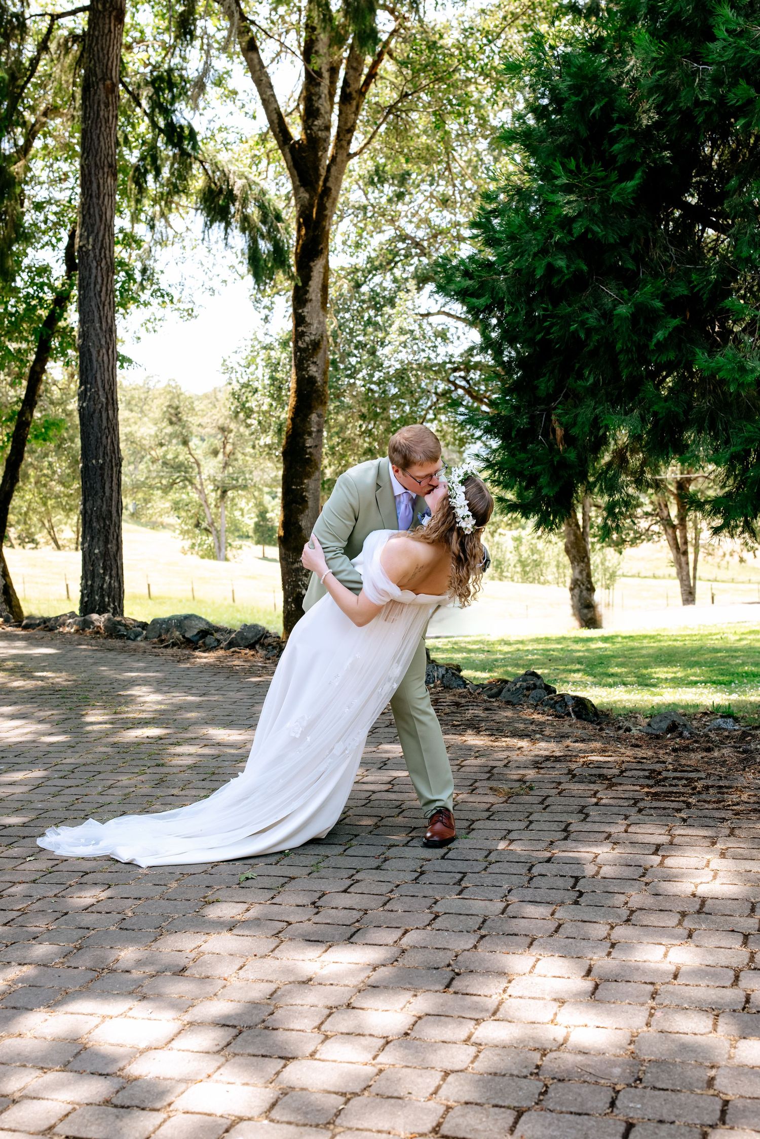 Groom dipping bride for a romantic kiss on a brick pathway surrounded by lush green trees at an outdoor wedding.
