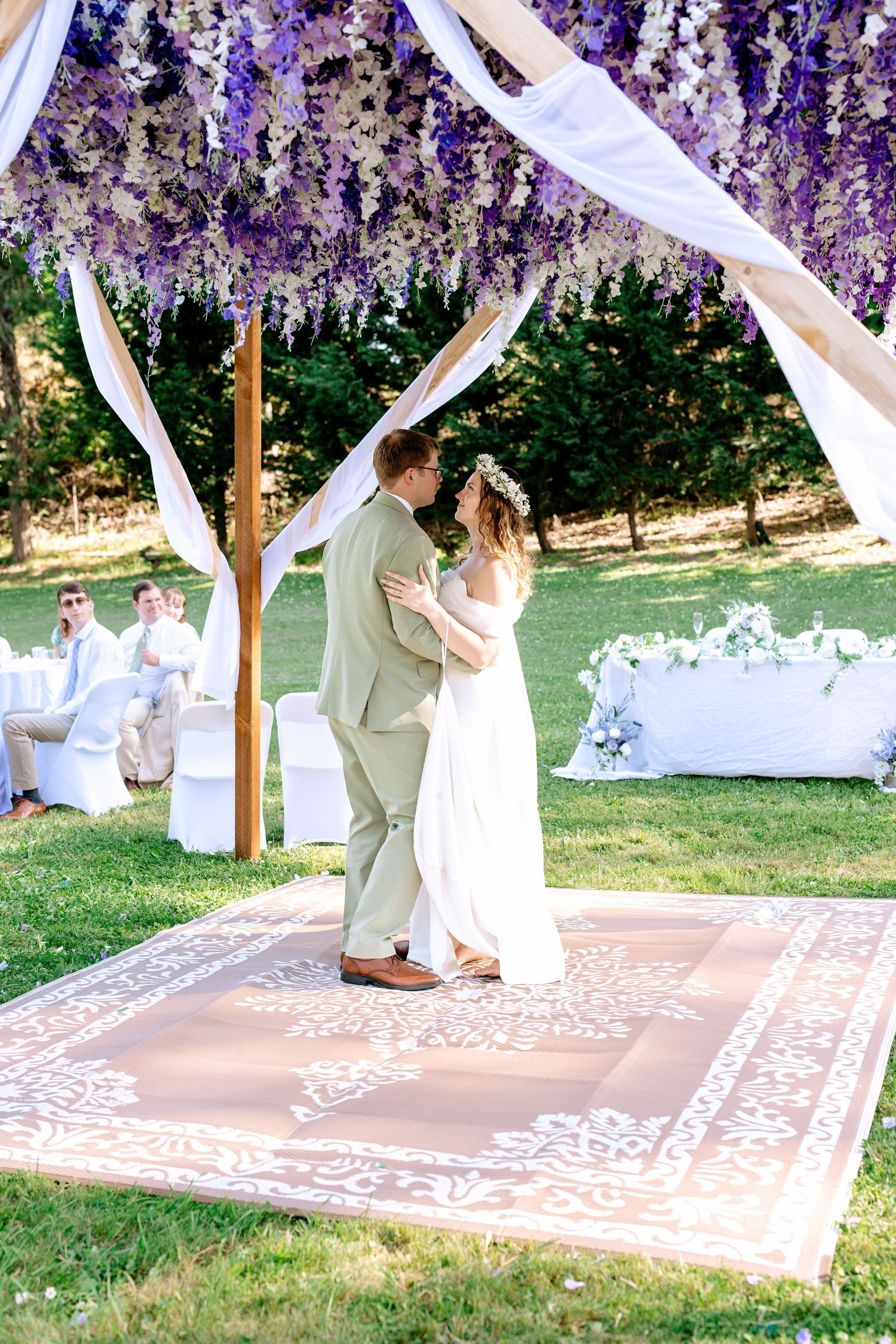 Bride and groom share first dance under stunning purple wisteria floral canopy at outdoor wedding reception.