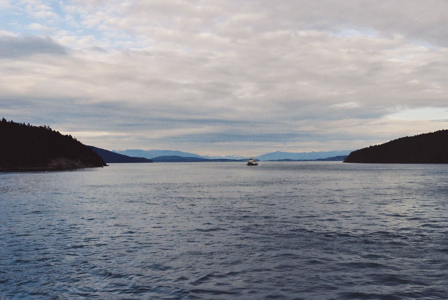 Calm blue ocean channel with small boat between forested hills under dramatic cloudy sky.