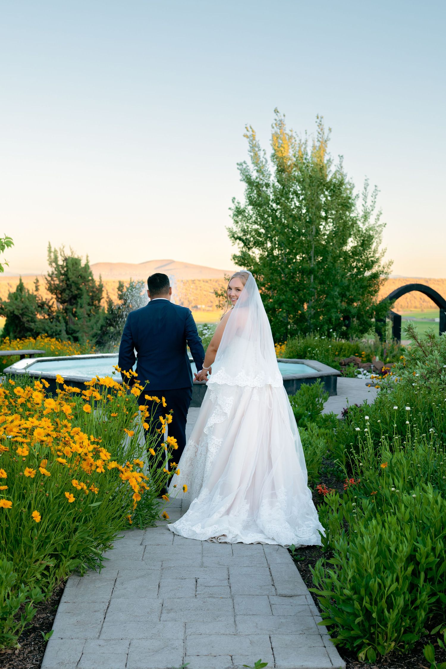 Bride and groom walking hand in hand through a garden path at sunset with mountain views.