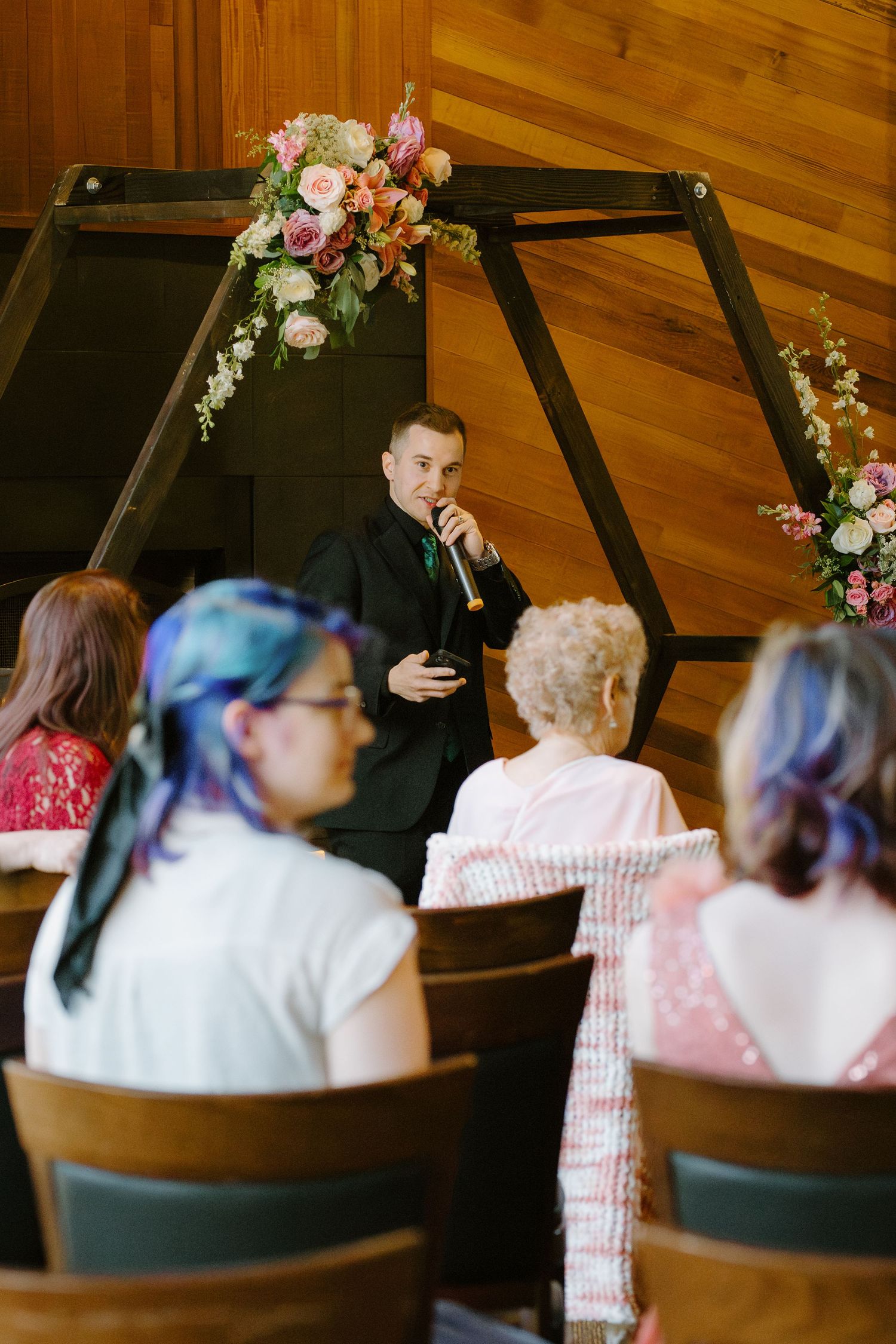 Officiant speaks at a wedding ceremony beneath a wooden arch adorned with pink and peach floral arrangements.