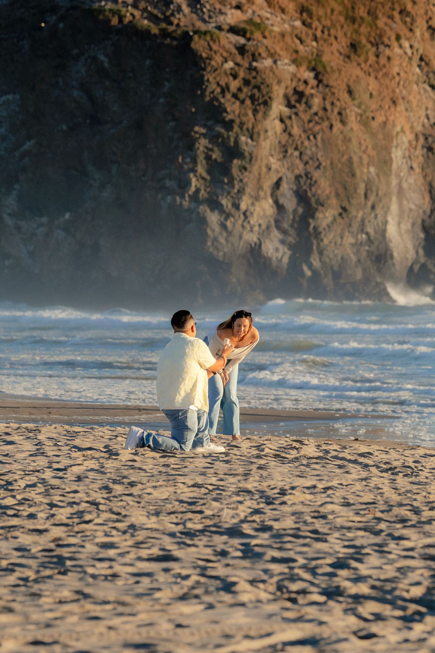 Man proposes on one knee to woman on sandy beach with ocean waves and rocky cliffs in background.