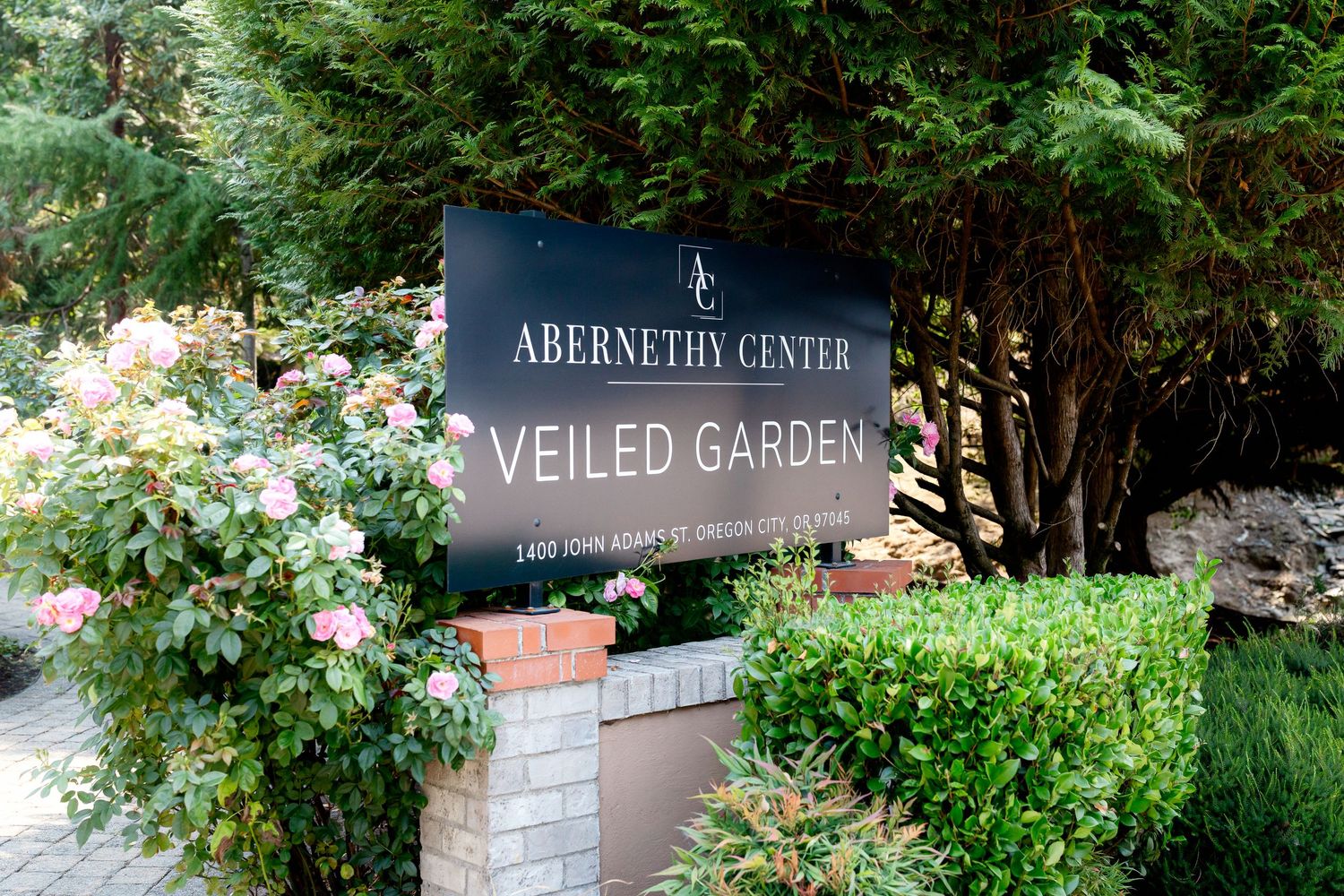 Sign for Abernethy Center Veiled Garden venue surrounded by lush greenery and pink roses in Oregon.