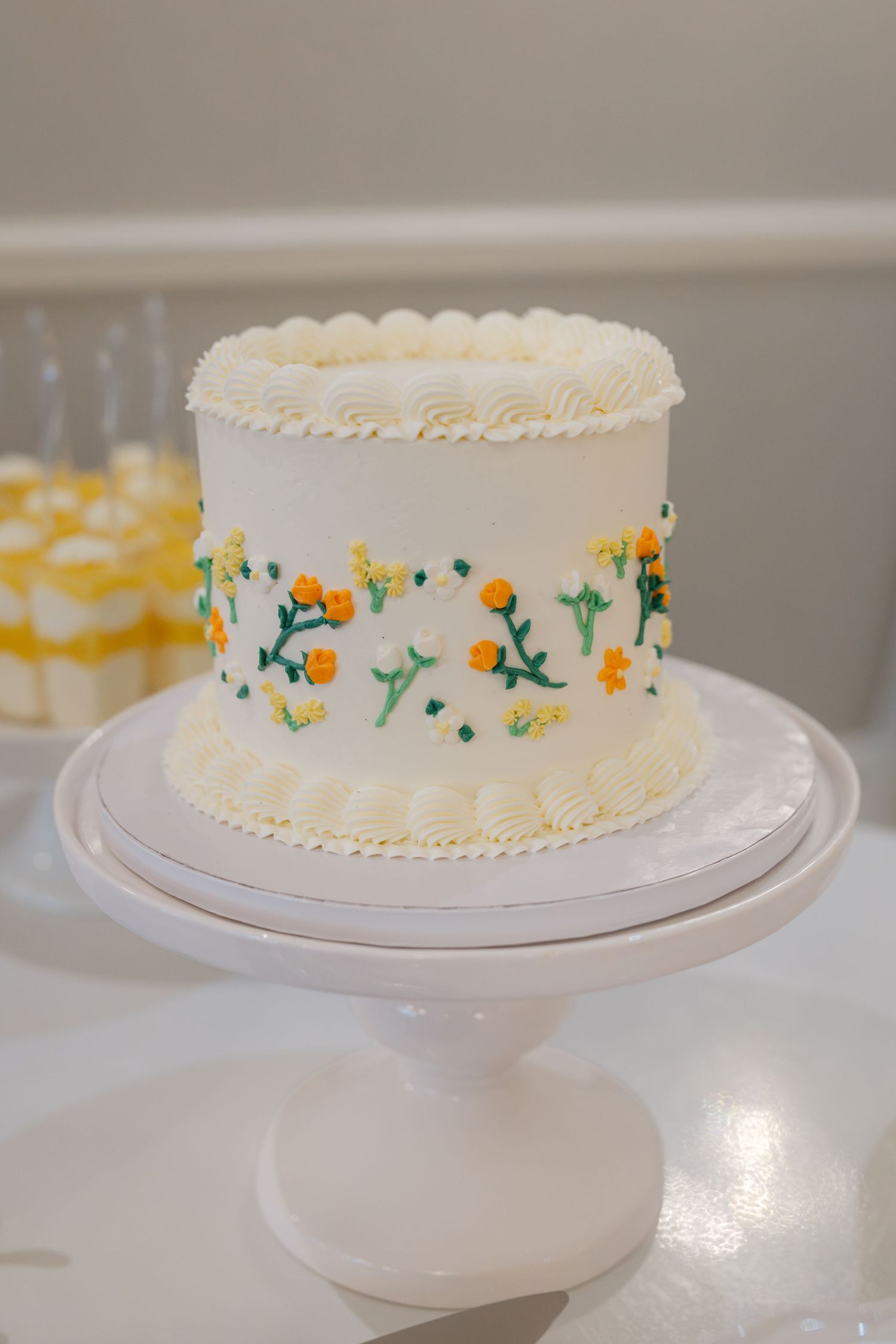 White wedding cake with yellow floral and green leaf piped decorations on a white pedestal stand.
