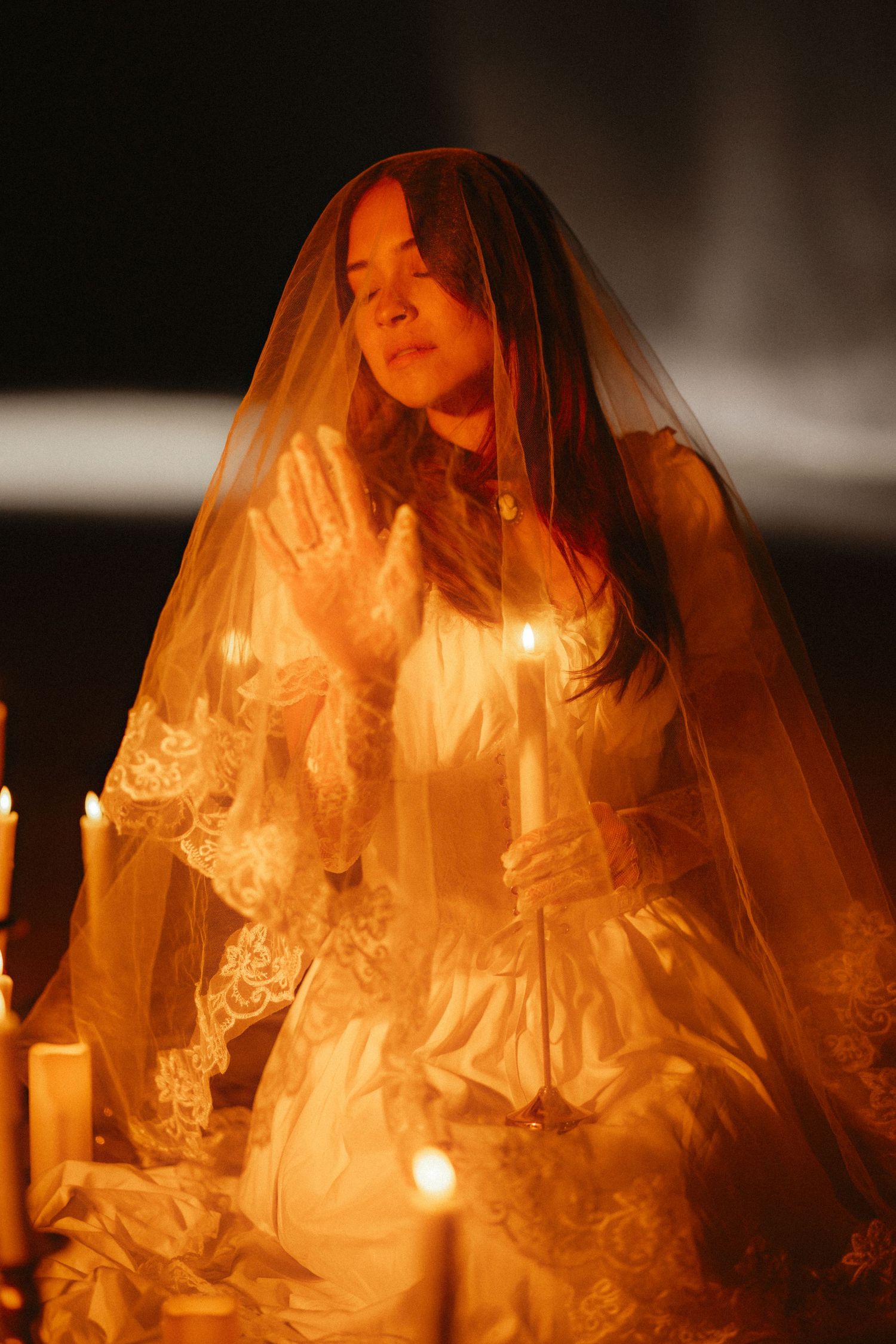 A figure in a vintage lace wedding veil poses dramatically in candlelight creating a moody, ethereal atmosphere.