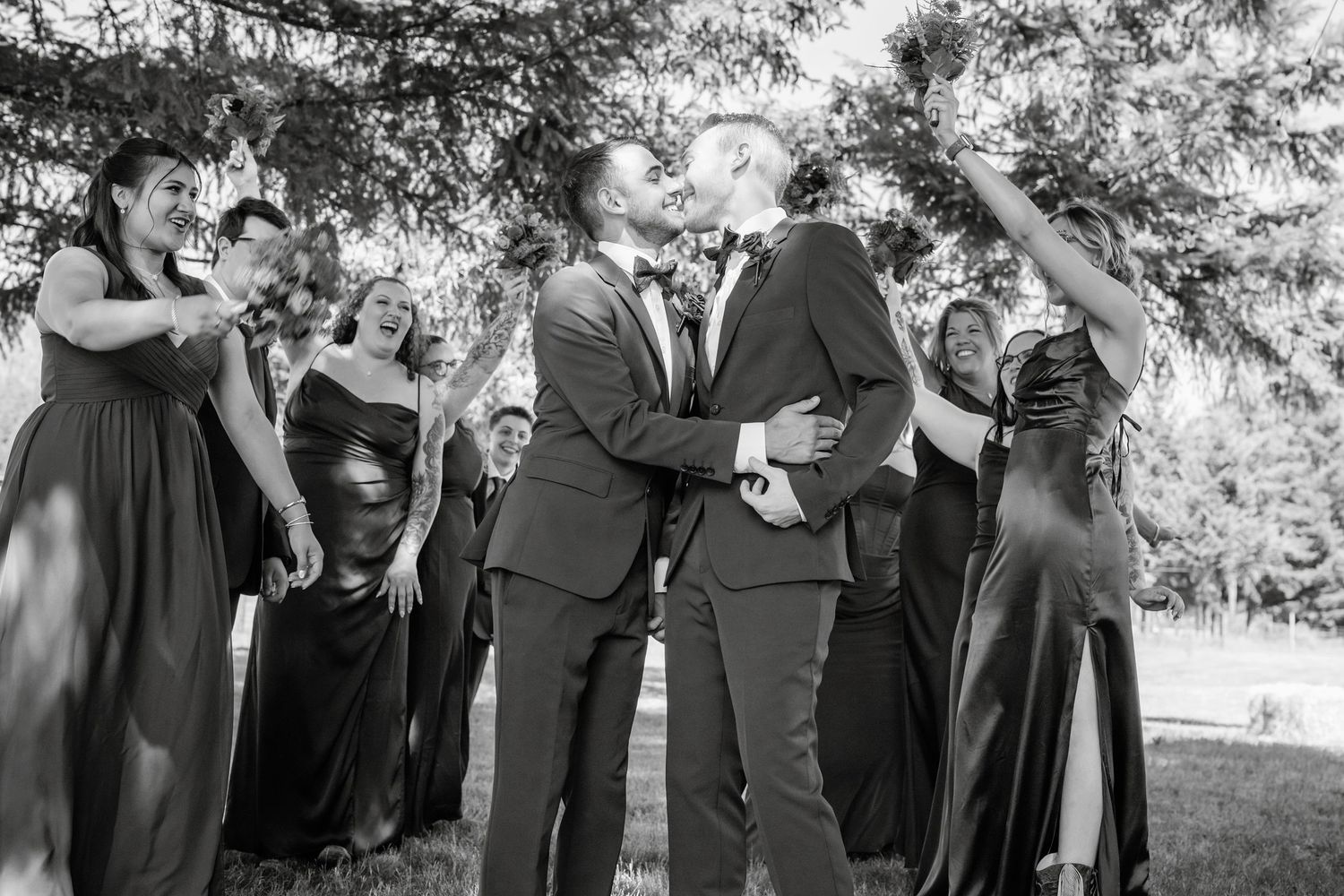 Black and white photo of two grooms kissing surrounded by cheering wedding party.