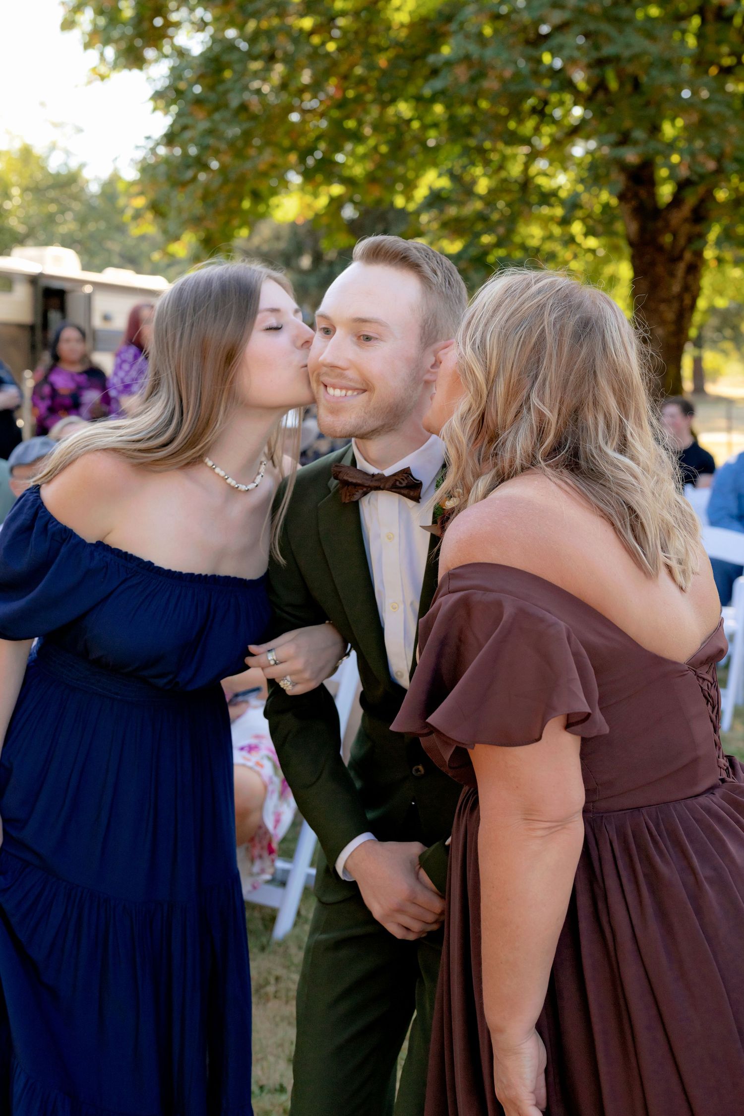Two women in blue and mauve dresses kiss a smiling groom in a green suit on the cheeks at an outdoor wedding.