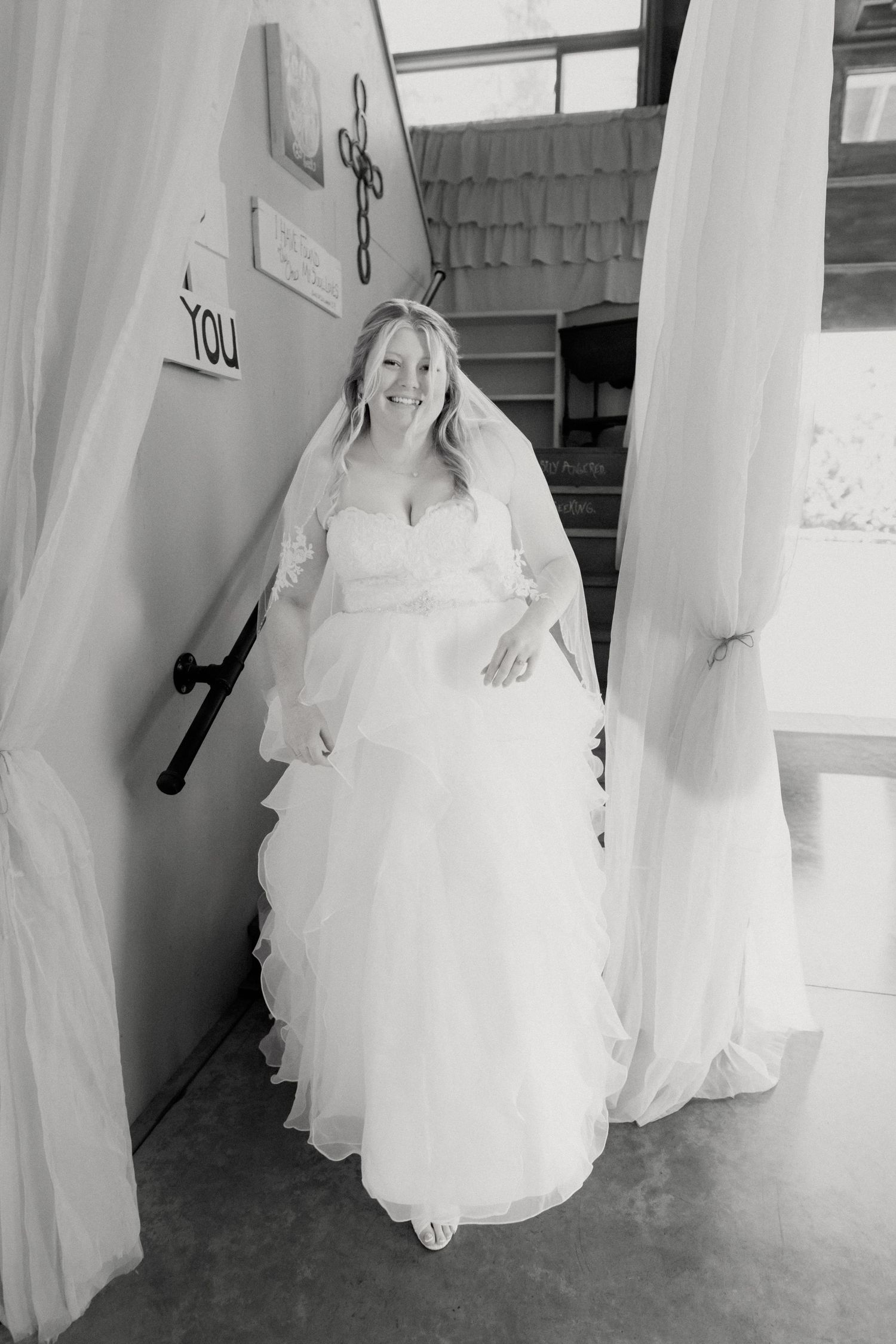 Smiling bride in white wedding gown and veil standing in doorway framed by white curtains, black and white photo.