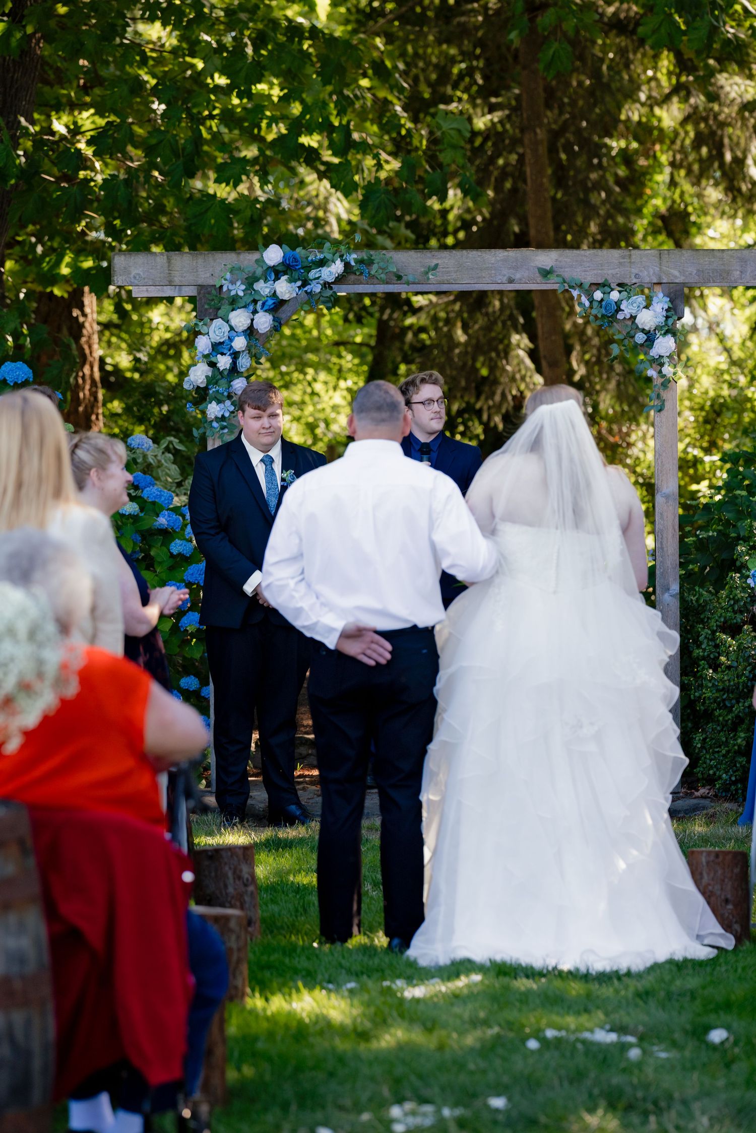 Bride and groom stand at floral arch during outdoor wedding ceremony, officiant and guests gathered in garden setting.
