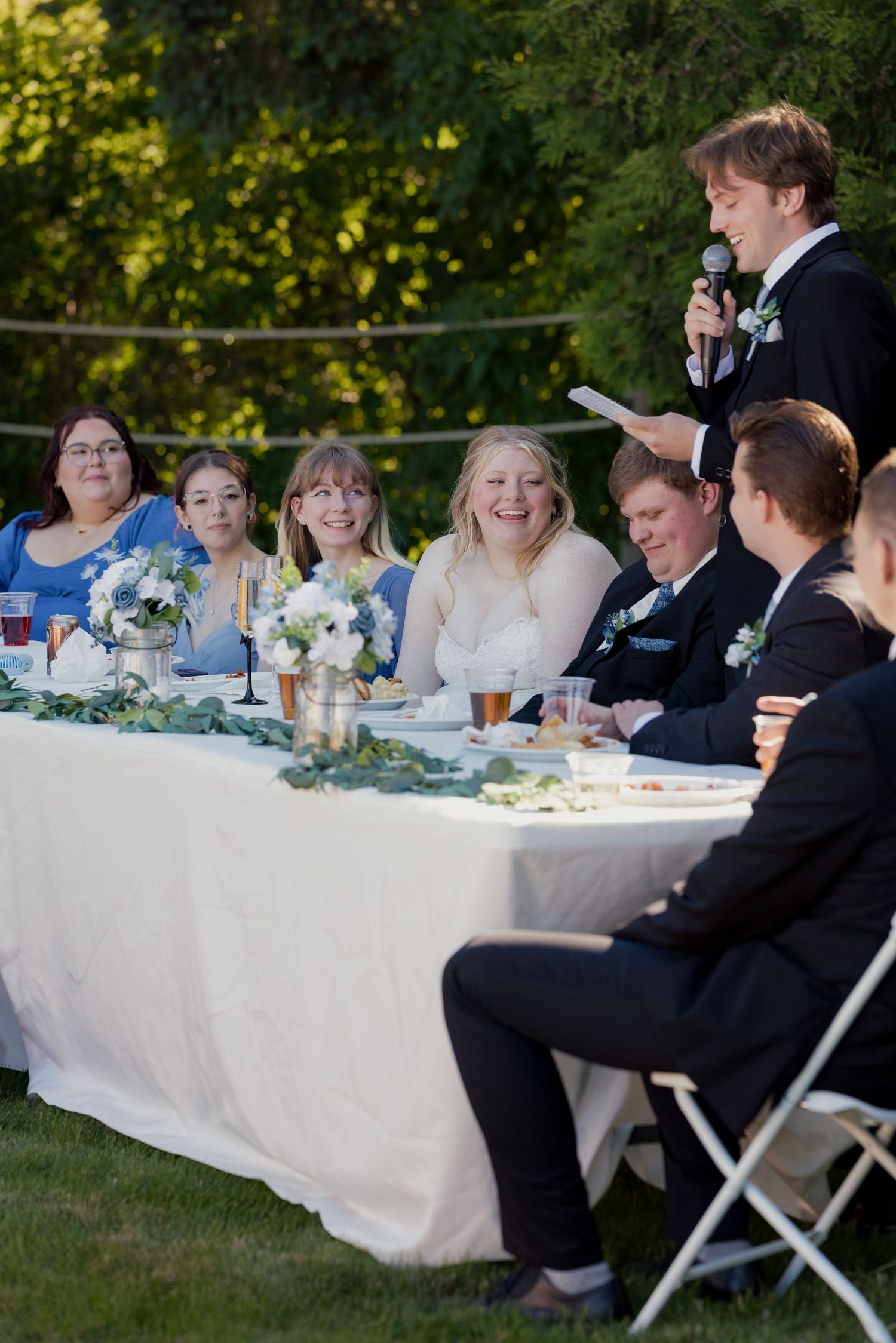 Wedding party seated at outdoor reception table while groomsman delivers speech into microphone.