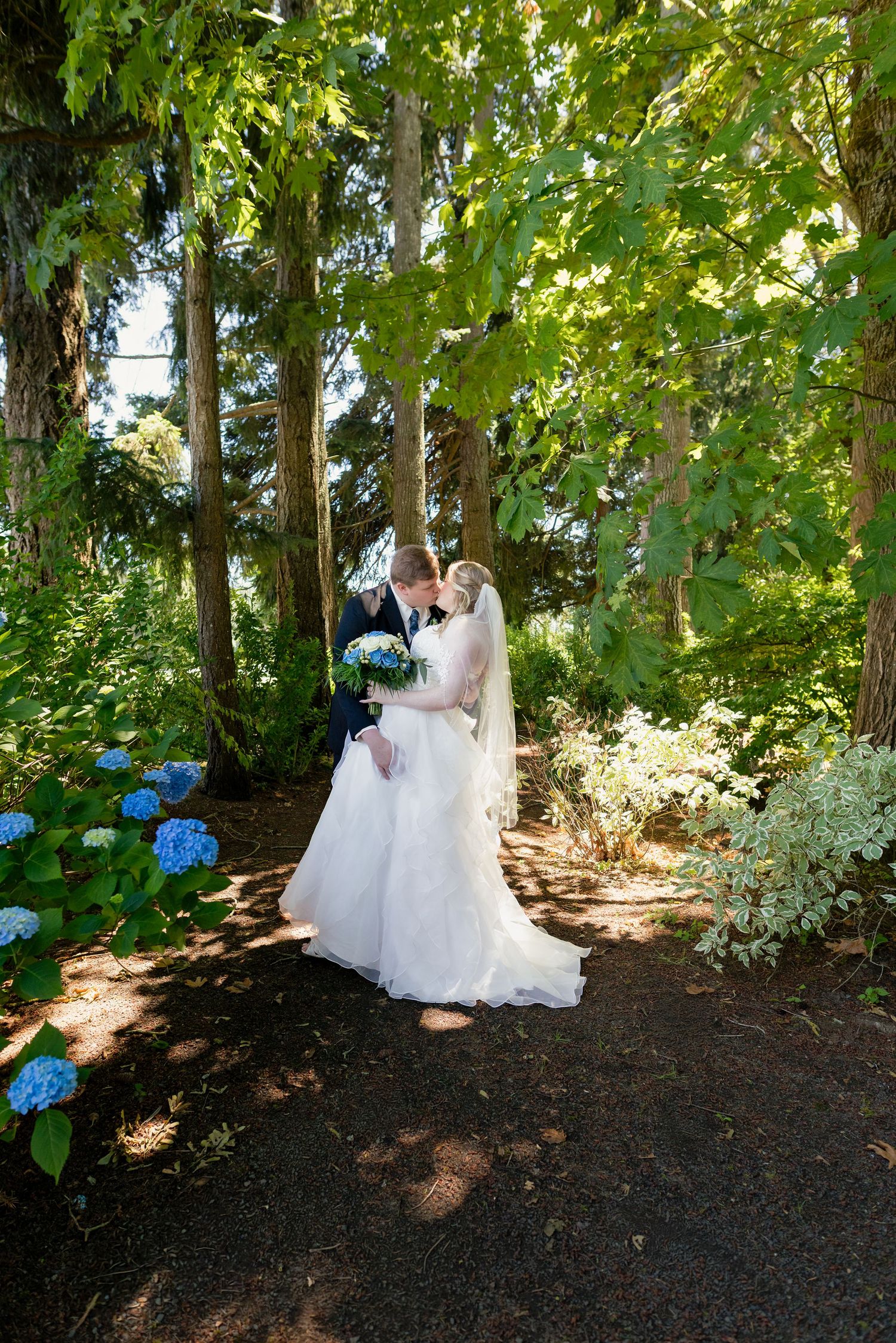 Bride and groom share a kiss on a garden path surrounded by lush green trees and blue hydrangeas.