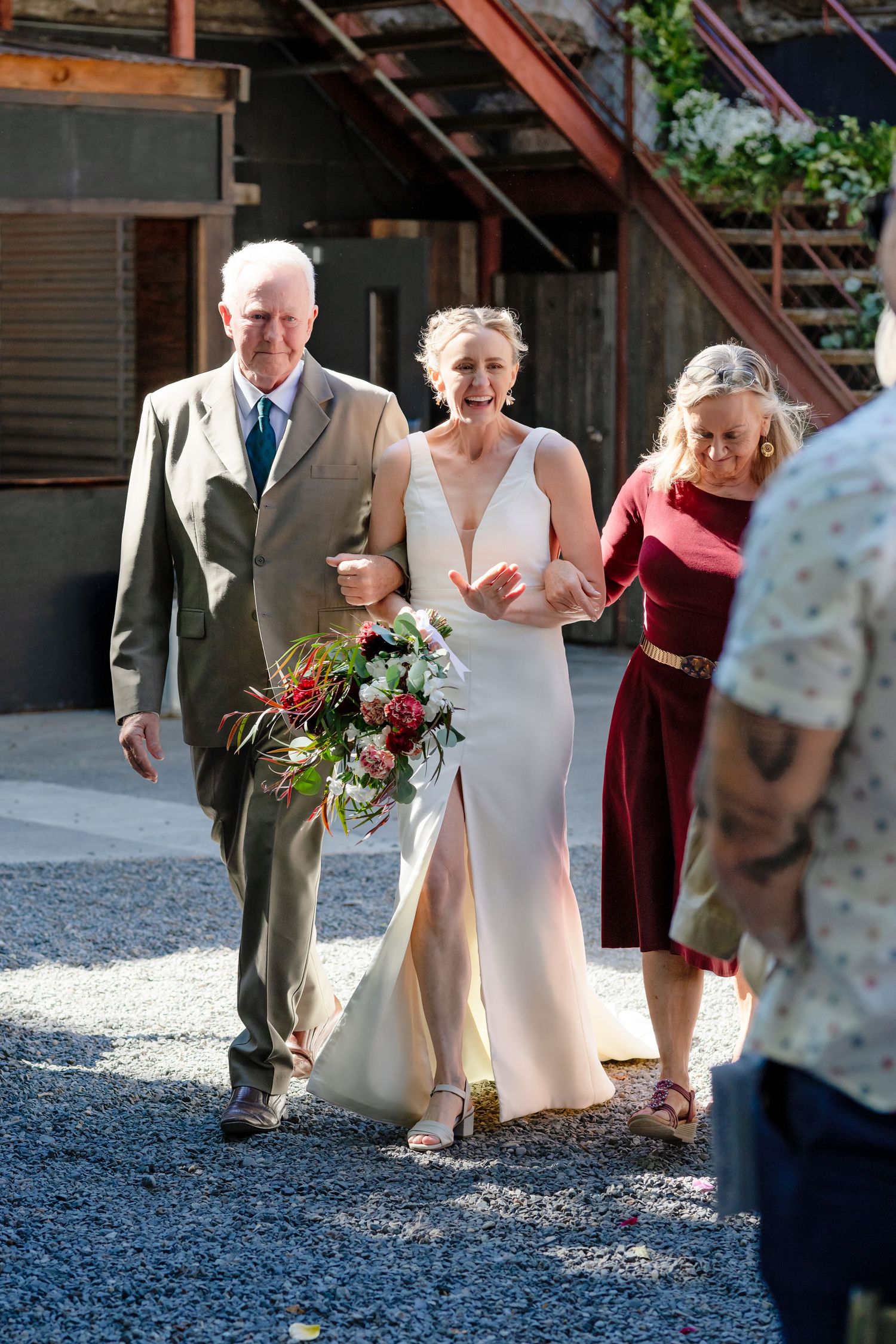 Radiant bride walks down the aisle escorted by parents at sunny outdoor wedding venue.