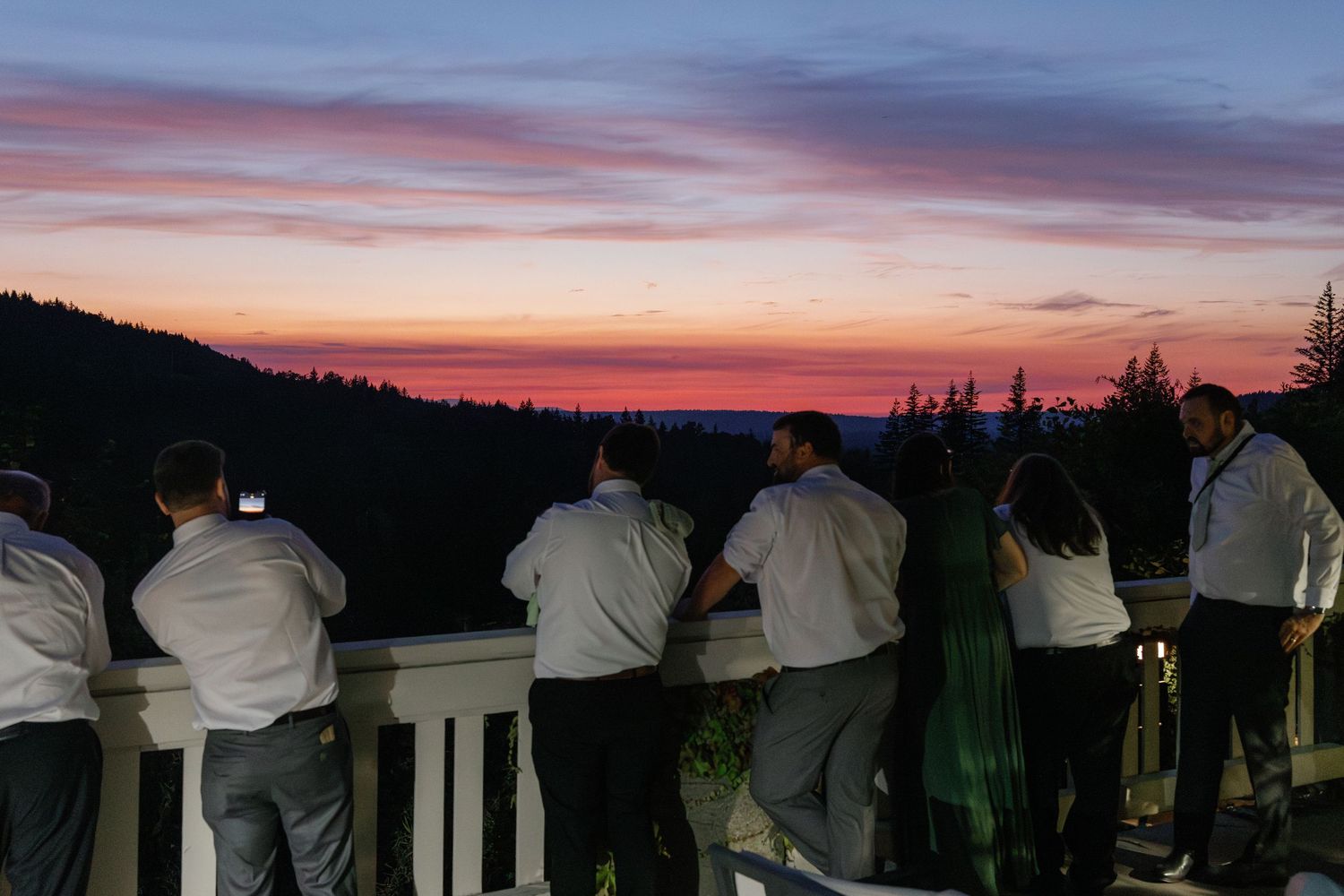 A series of people lined up along a railing at dusk watching a colorful sunset over a dark landscape.