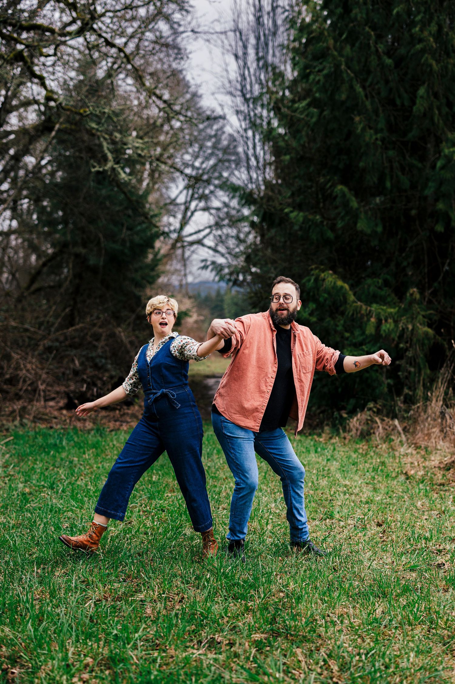 A cheerful couple strikes playful poses while standing in a grassy field surrounded by evergreen trees.