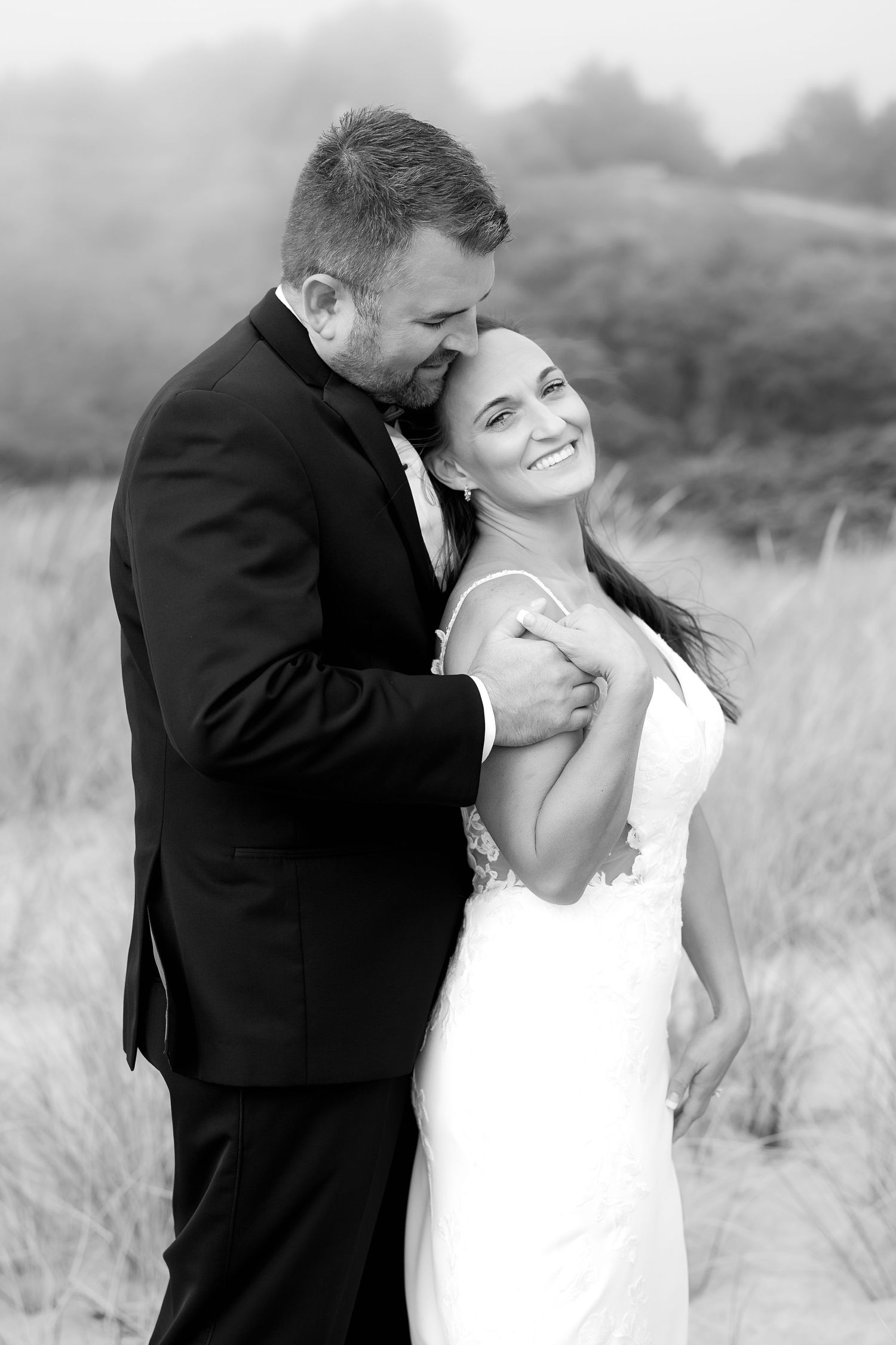 Black and white romantic portrait of newlyweds embracing in coastal grass field.