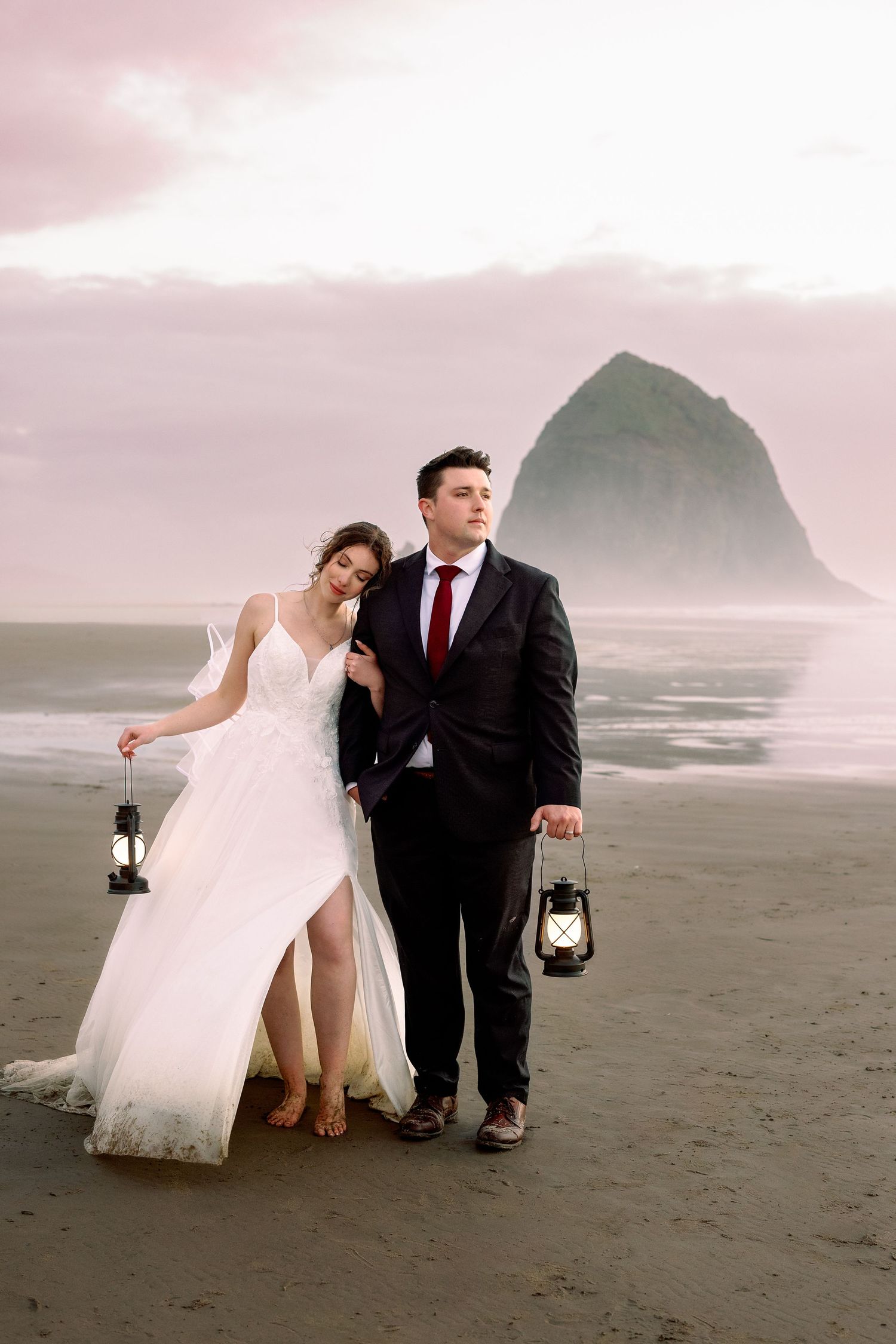 Newlyweds pose with vintage lanterns on a moody beach with coastal rock formations in the distance.