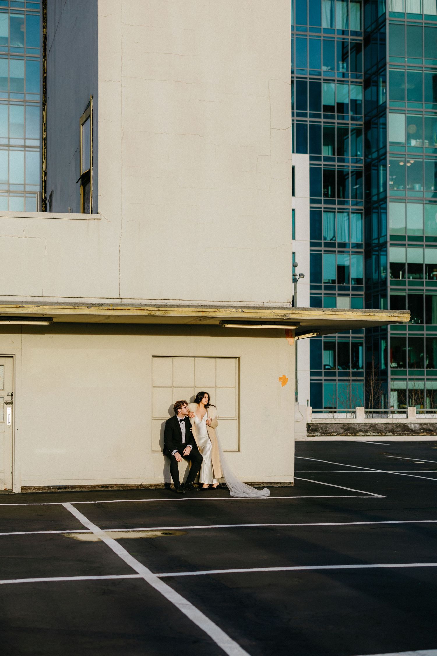 A couple poses for wedding photos in an urban setting with glass buildings and empty parking lot in the background.