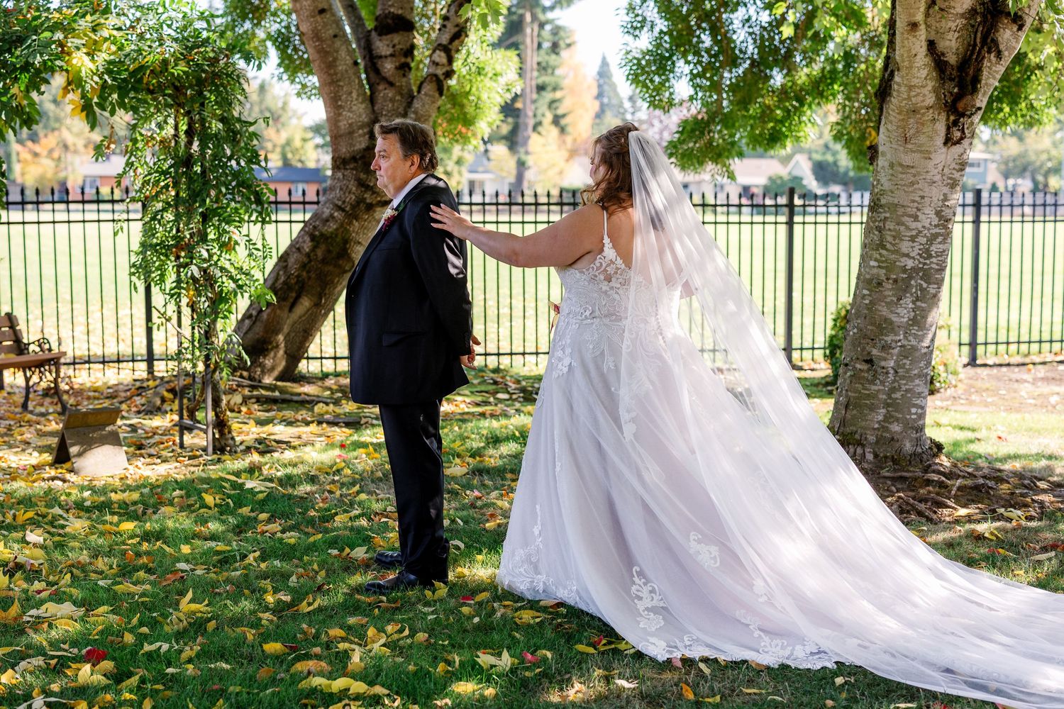 A couple in wedding attire stands in a park setting with autumn leaves scattered on the ground under tall trees.