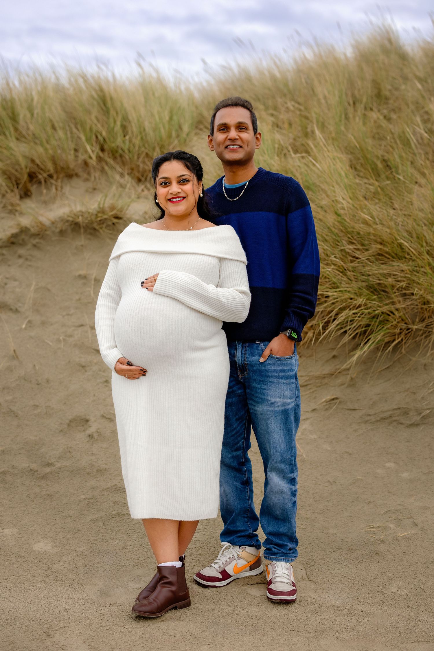 People in casual attire pose playfully on a sandy beach with tall grass in the background.
