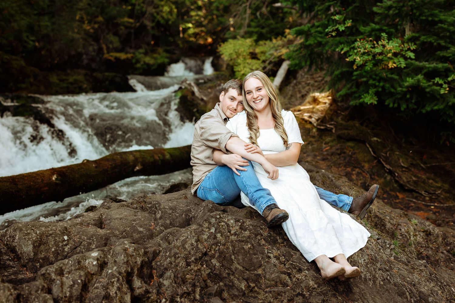 A couple sits together on mossy rocks near a cascading waterfall in a lush forest setting.