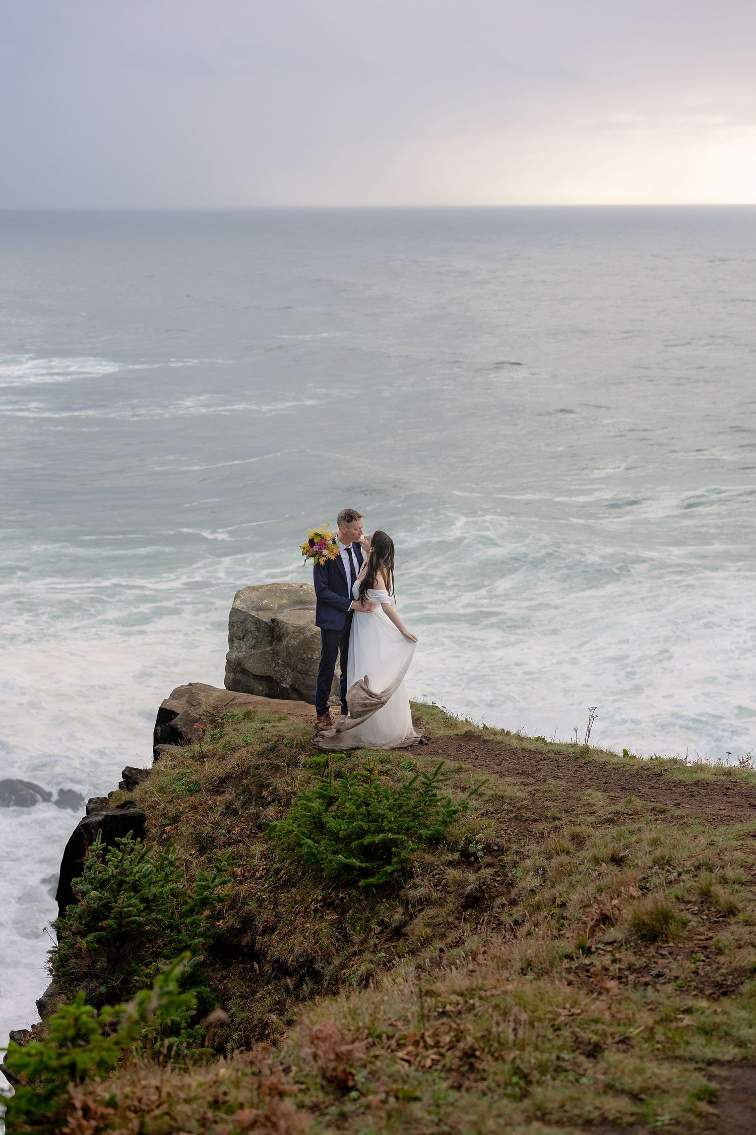 A couple stands on a dramatic coastal cliff overlooking the misty ocean waves during a romantic sunset moment.