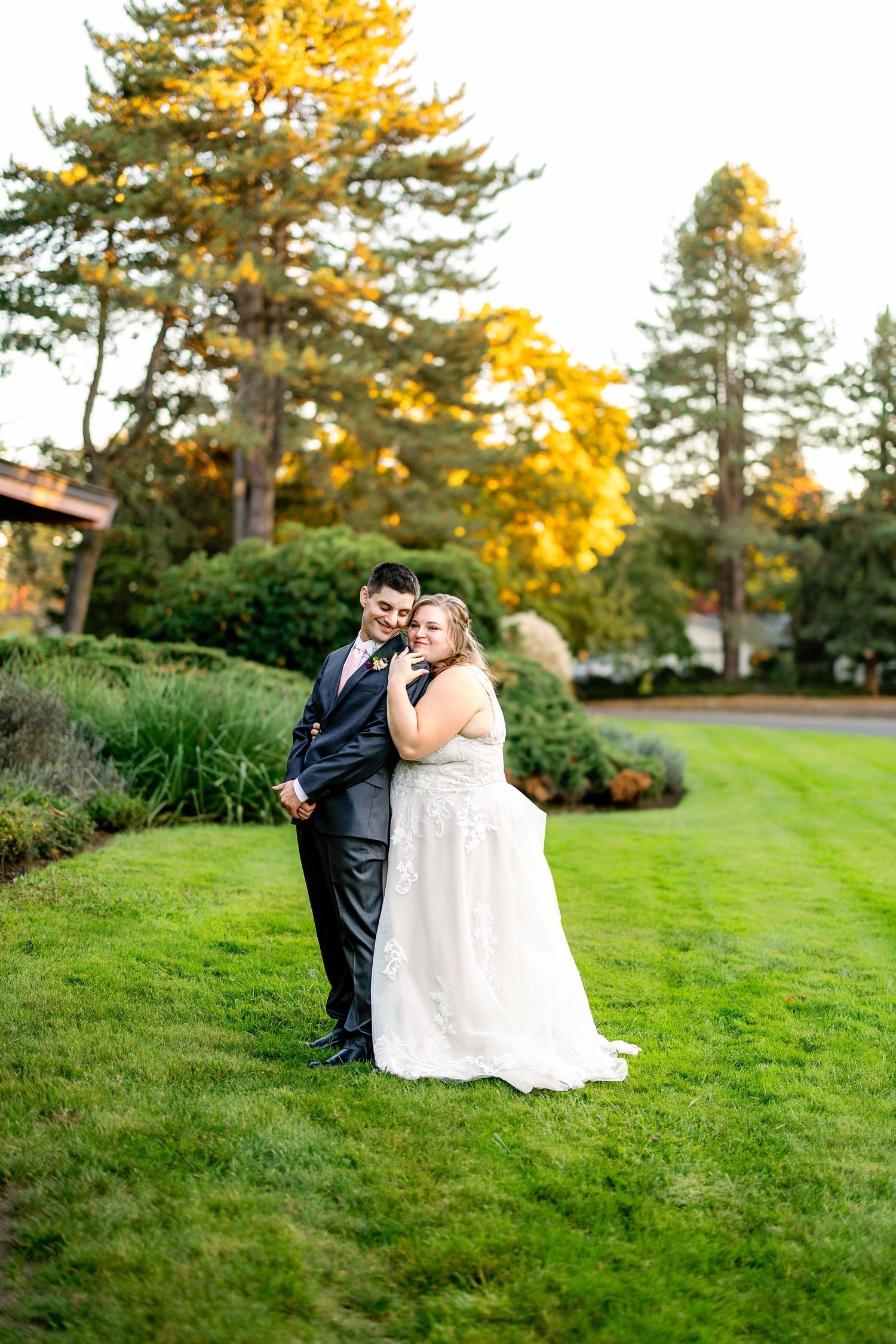 A couple poses for wedding photos on a lush lawn with autumn trees in the background.