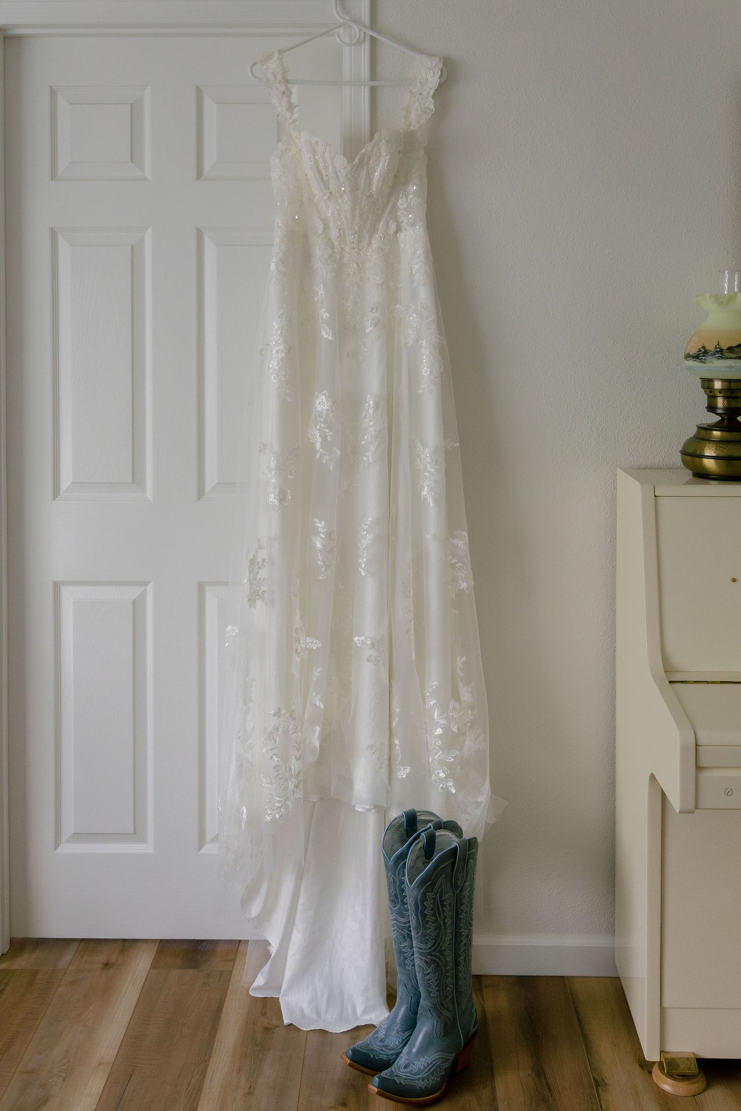 A white lace wedding dress hangs on a door with turquoise cowboy boots placed beneath it on hardwood flooring.