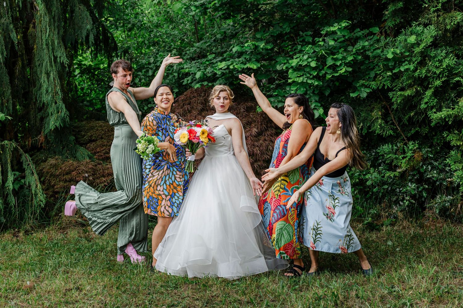 A group of wedding guests enjoy a playful moment together on a grassy hillside surrounded by lush green foliage.