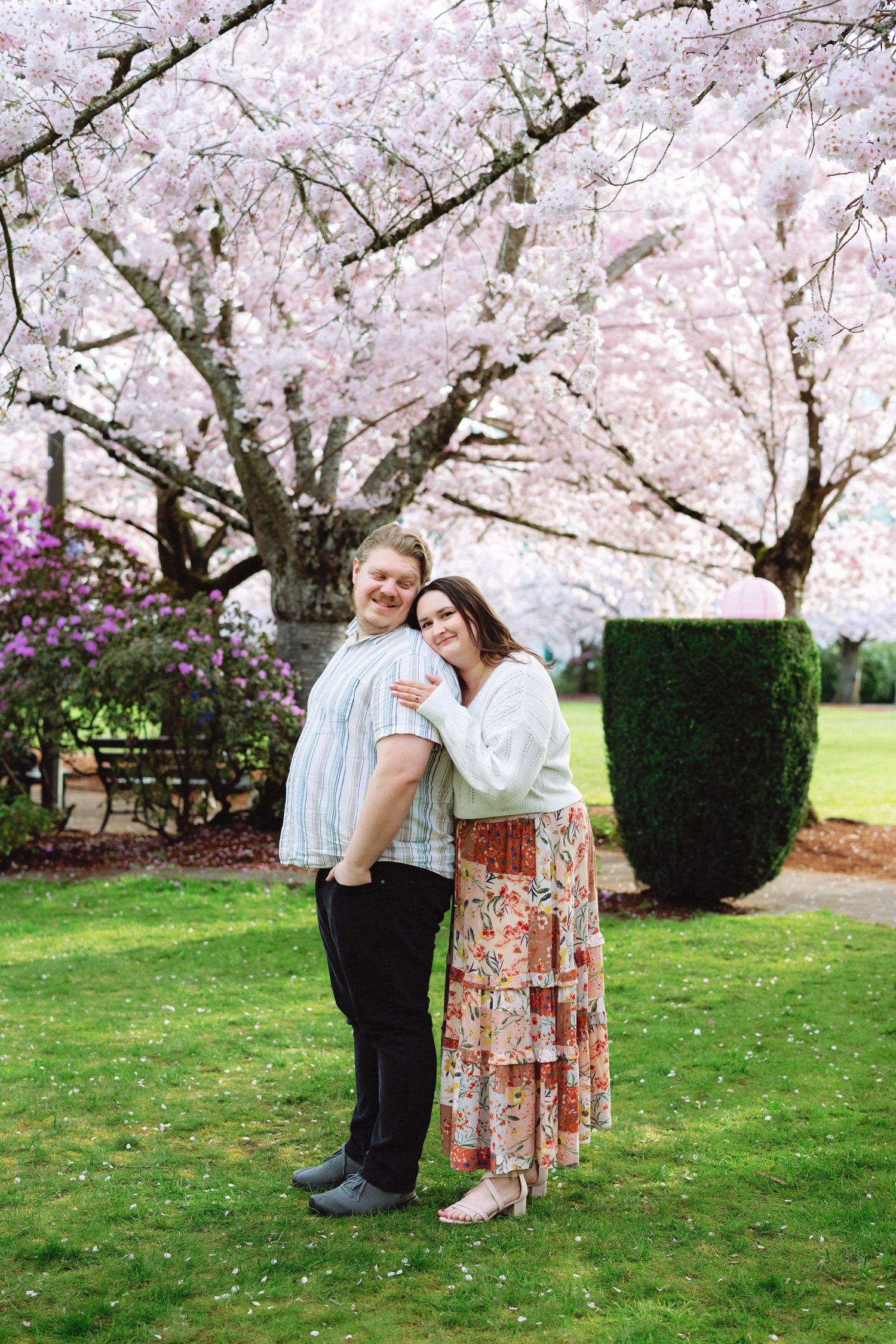 A romantic couple poses together under blooming cherry blossom trees in a spring garden setting.