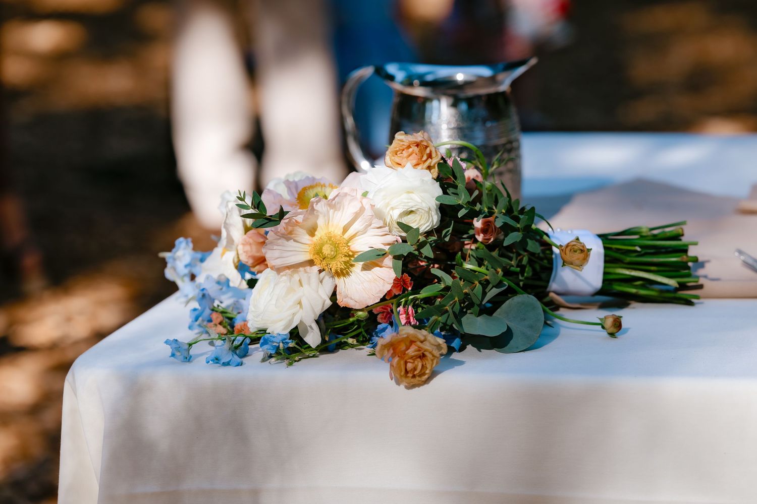 Elegant wedding bouquet with white peonies, blue flowers, and greenery on a white tablecloth outdoors.