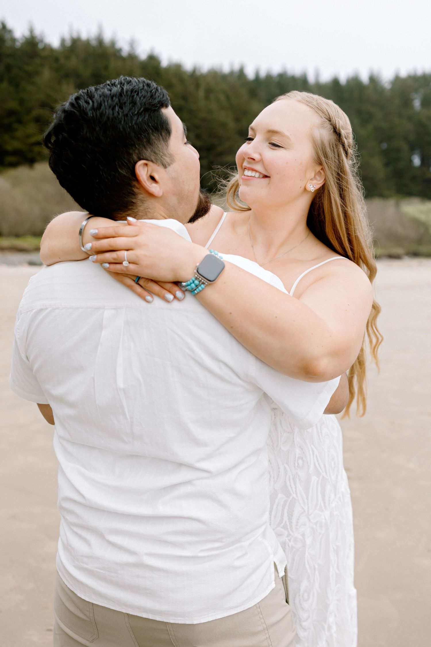 Woman in white dress smiling while embracing man in white shirt on a beach with evergreen trees in background.