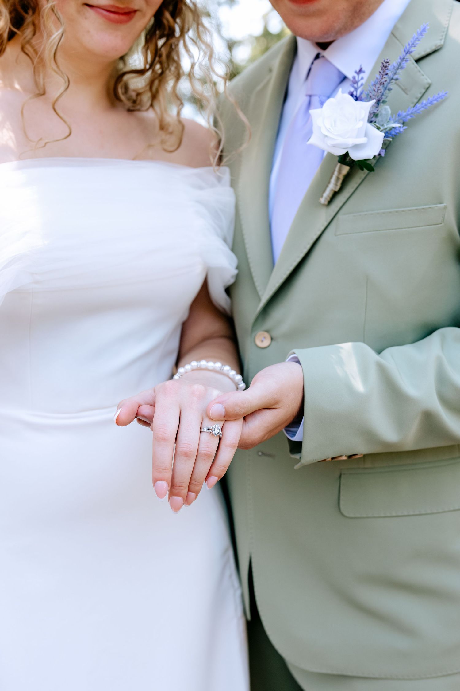 Bride and groom holding hands showing wedding ring and pearl bracelet, groom in sage suit with white boutonniere.