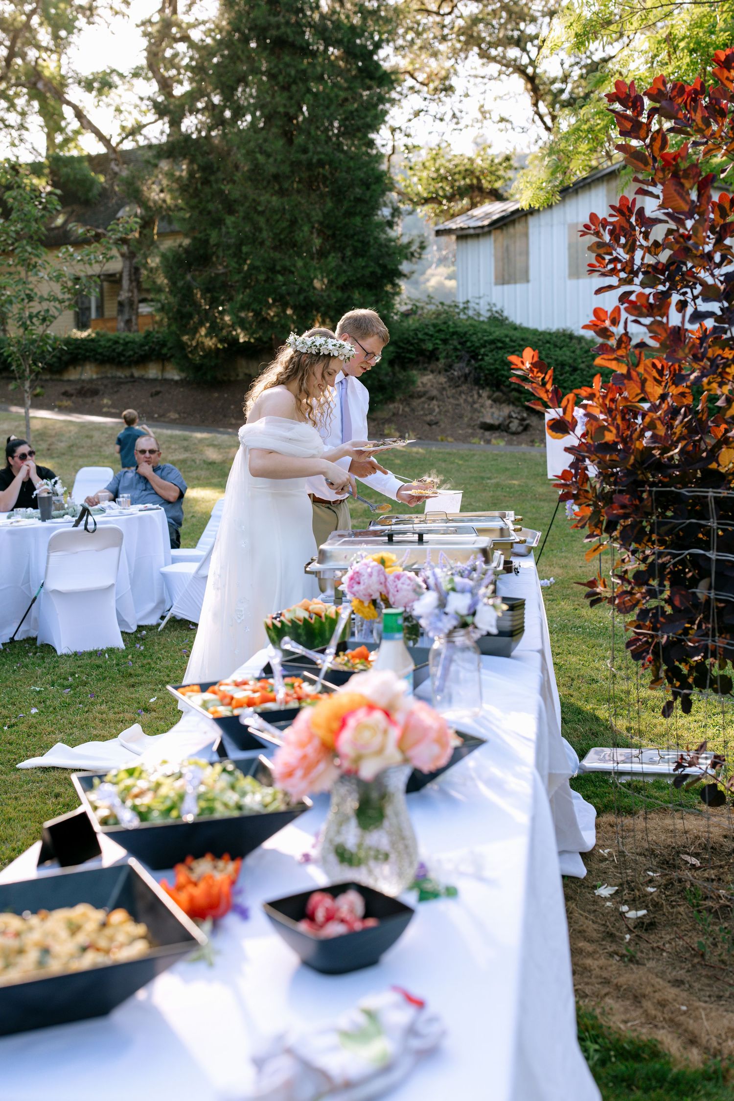 Newlyweds serve themselves at an elegant outdoor wedding buffet with floral arrangements.