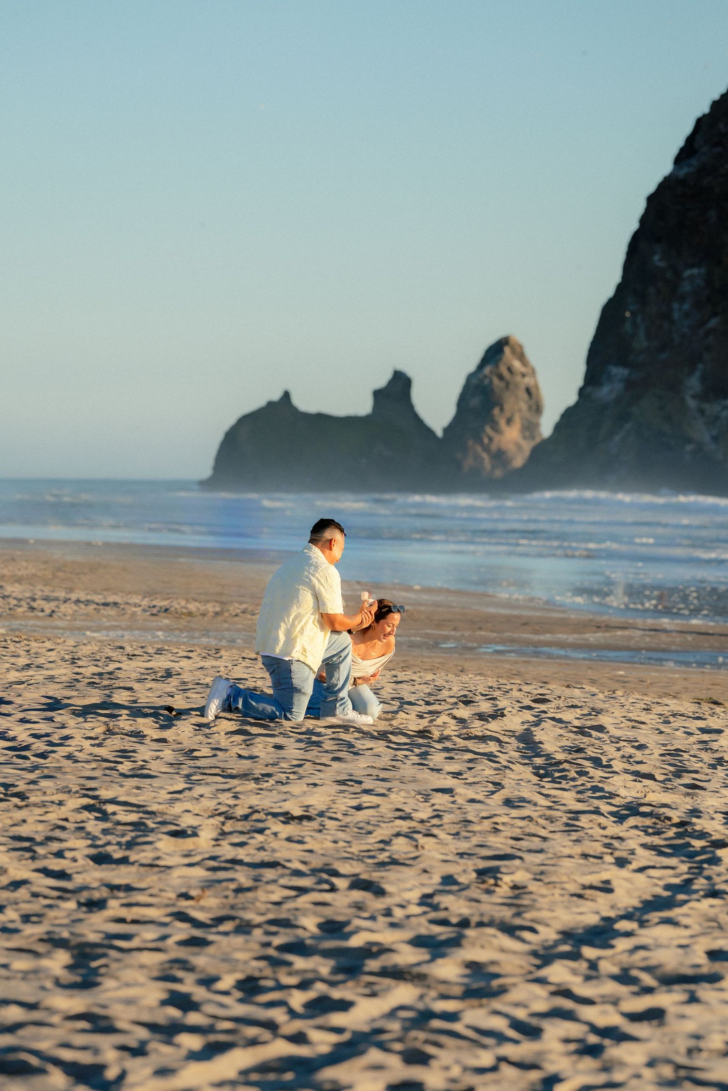 Man proposing on one knee to woman on Cannon Beach at sunset with sea stacks in background.