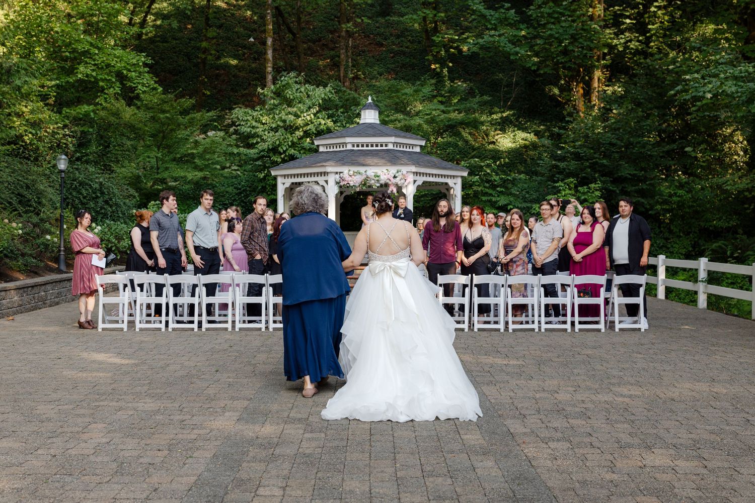 Bride walks toward outdoor gazebo wedding ceremony with guests seated in white chairs.