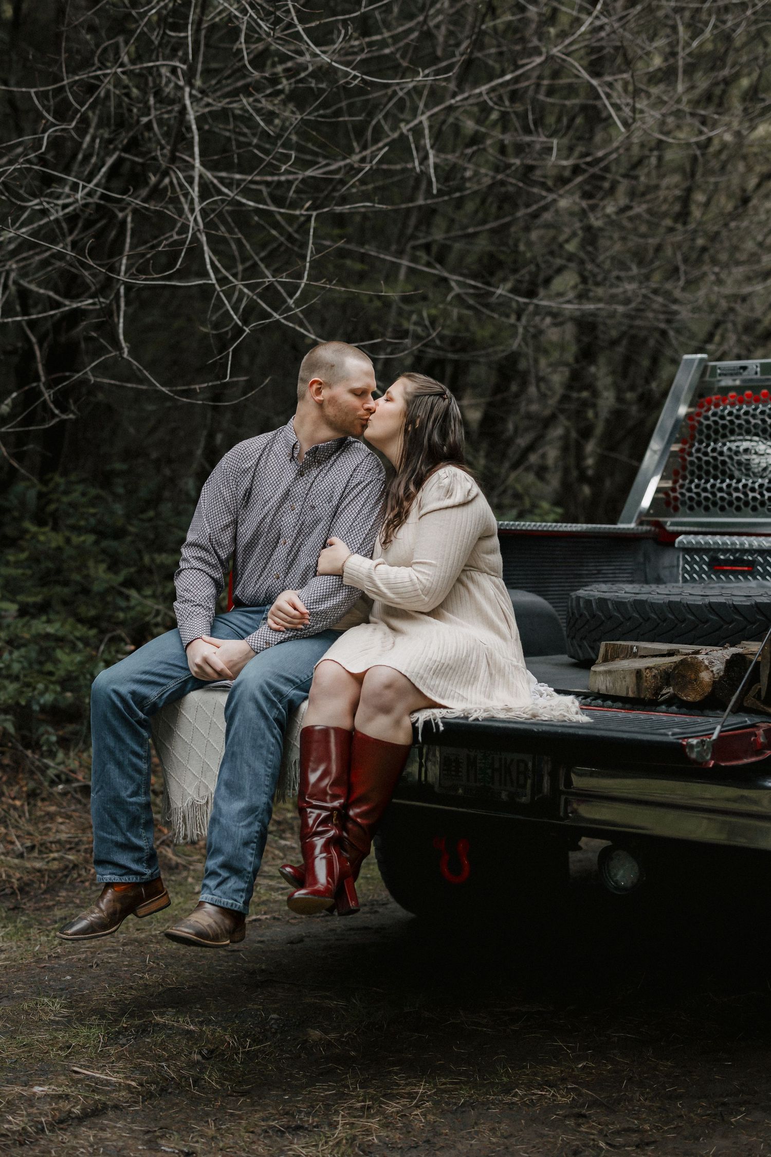 Couple sitting together on the back of a pickup truck in a wooded setting at dusk.