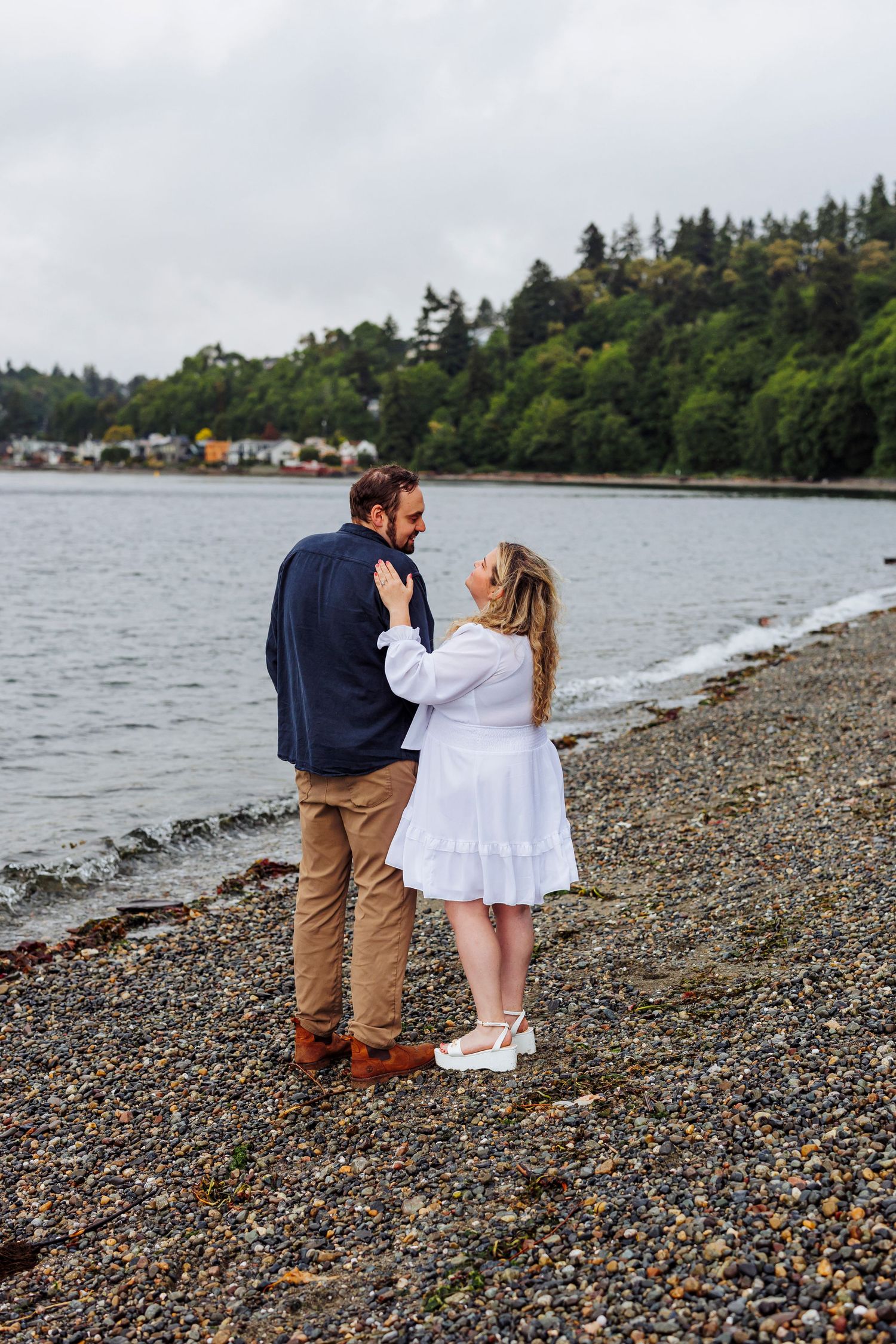 A couple embraces while standing on a rocky beach shore with evergreen trees and boats visible in the background.