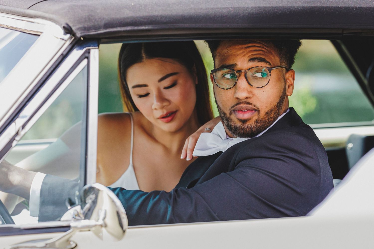 A couple dressed in formal attire sit together in a vintage car, with the driver playfully making a surprised expression.
