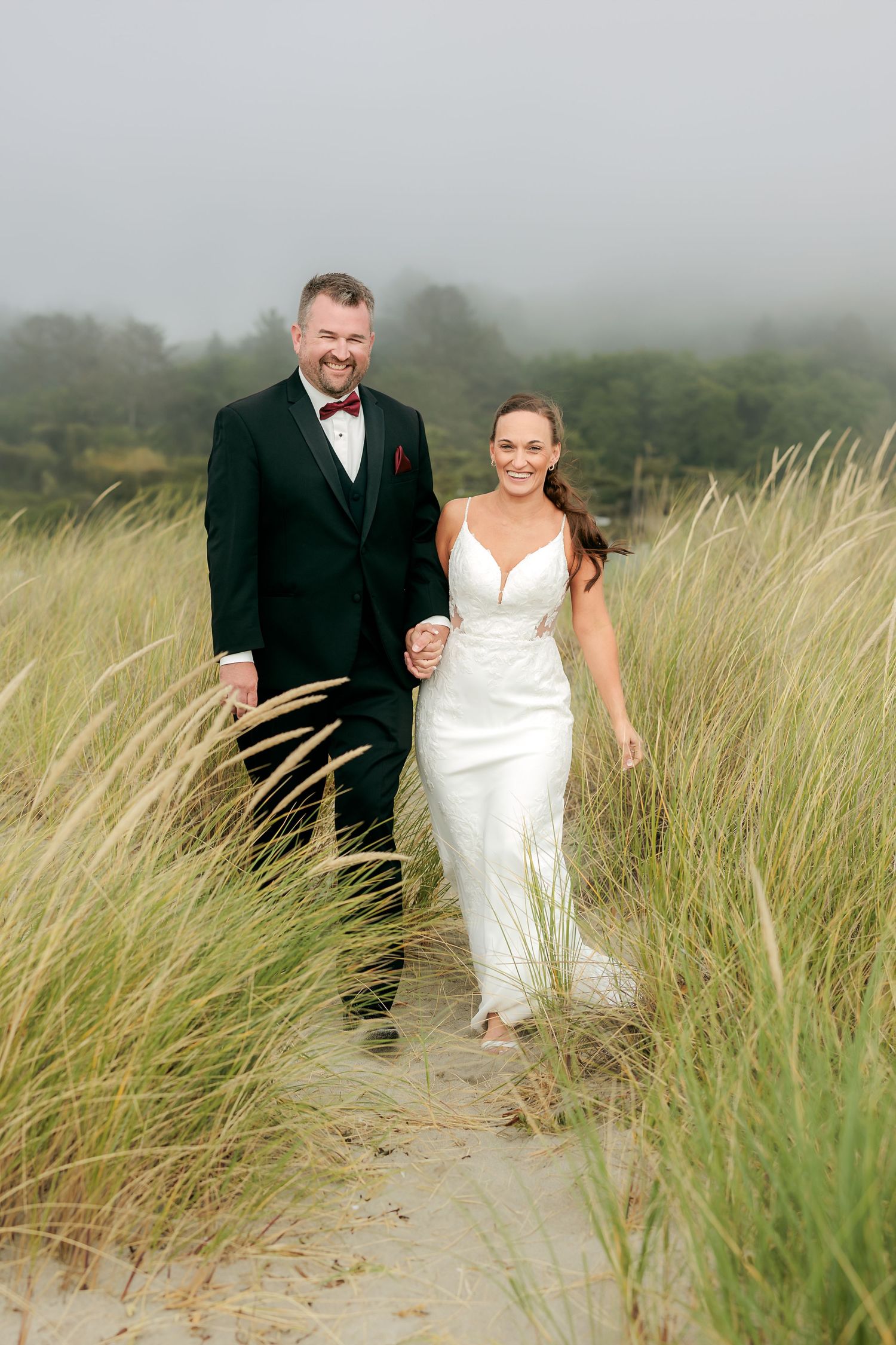 A couple walks together through coastal dunes on their wedding day in misty weather.