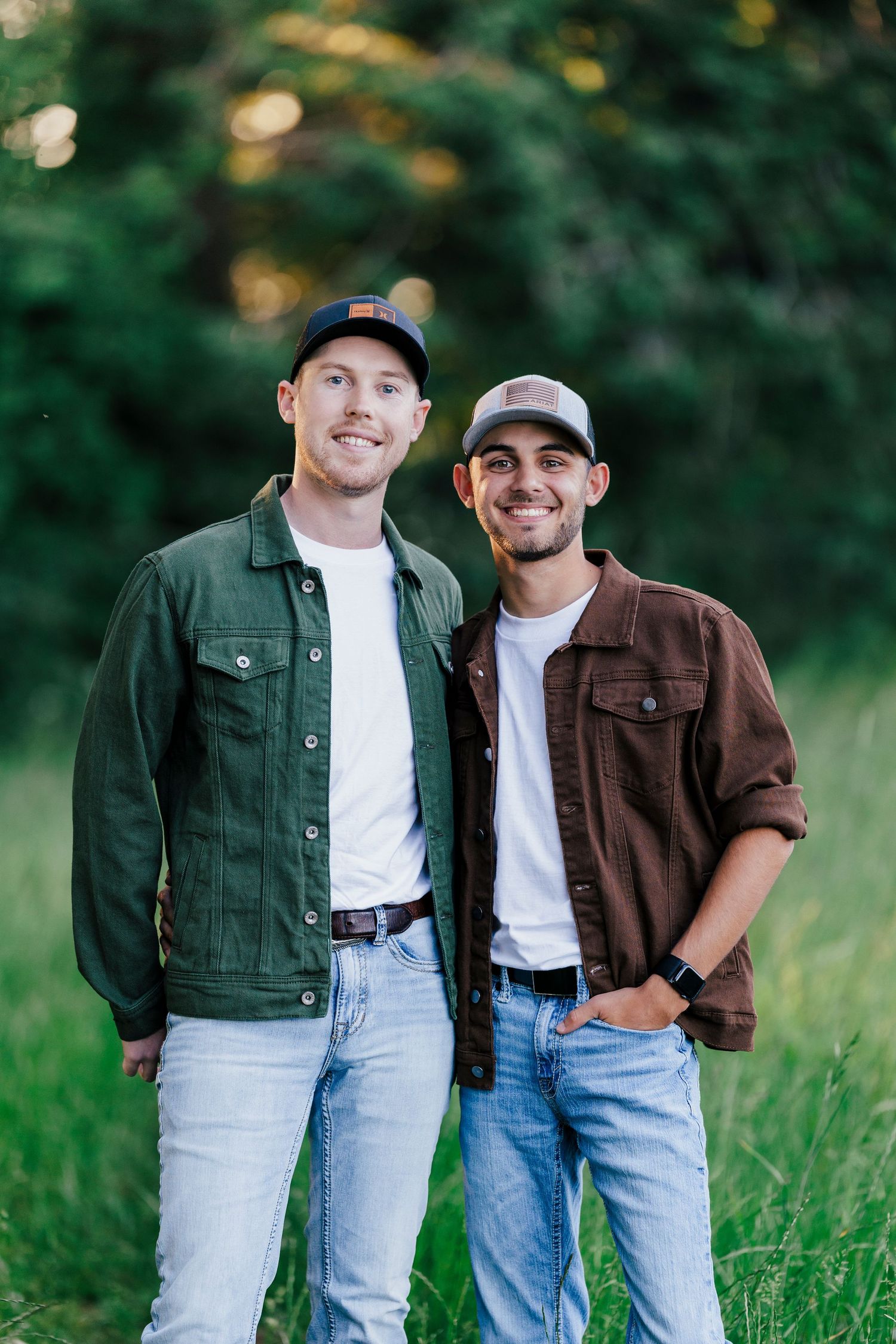 Two people in casual denim and jackets pose together in a grassy outdoor setting.