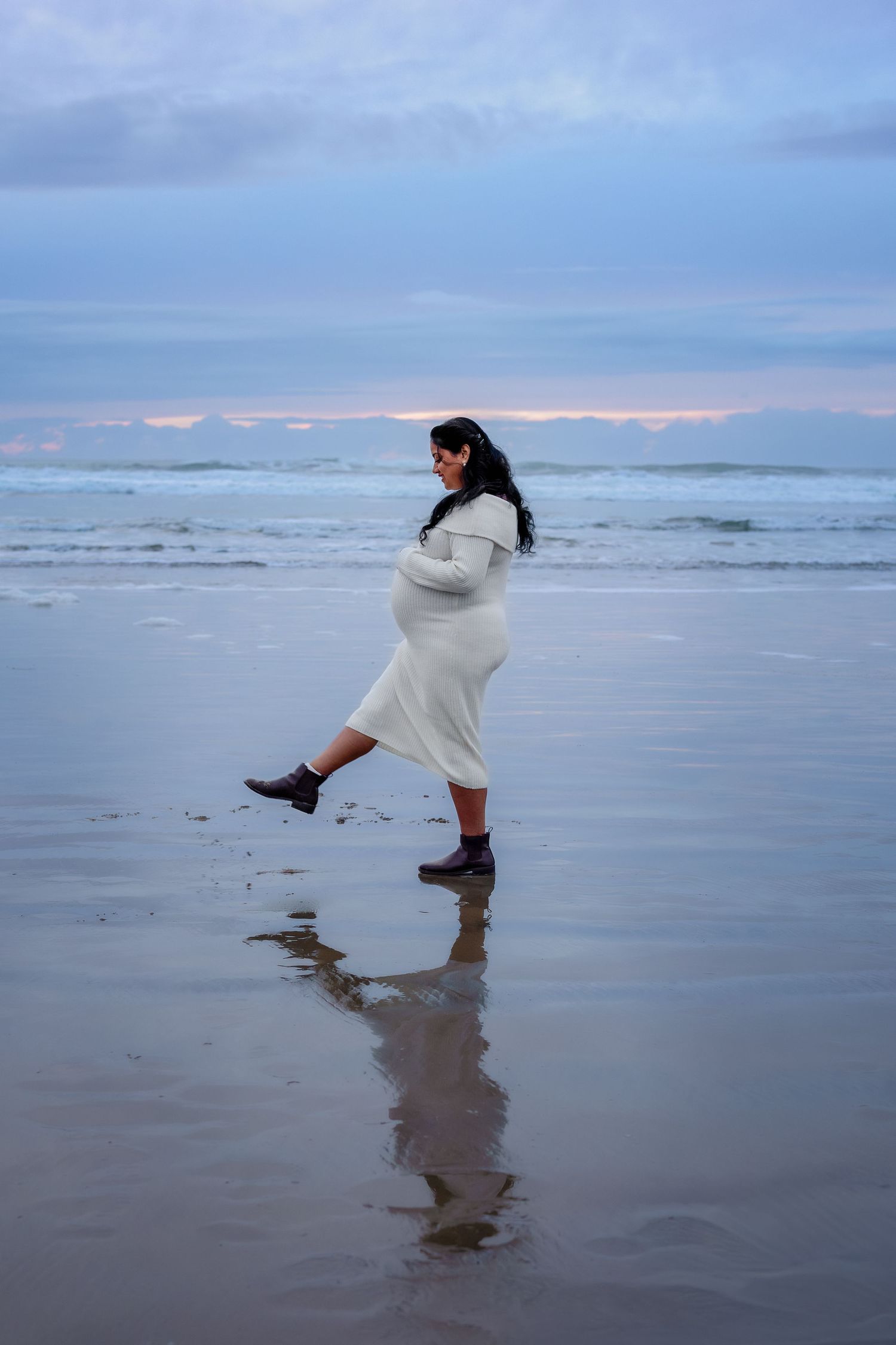 Someone wearing a white sweater playfully kicks water on a moody Pacific Northwest beach at dusk.