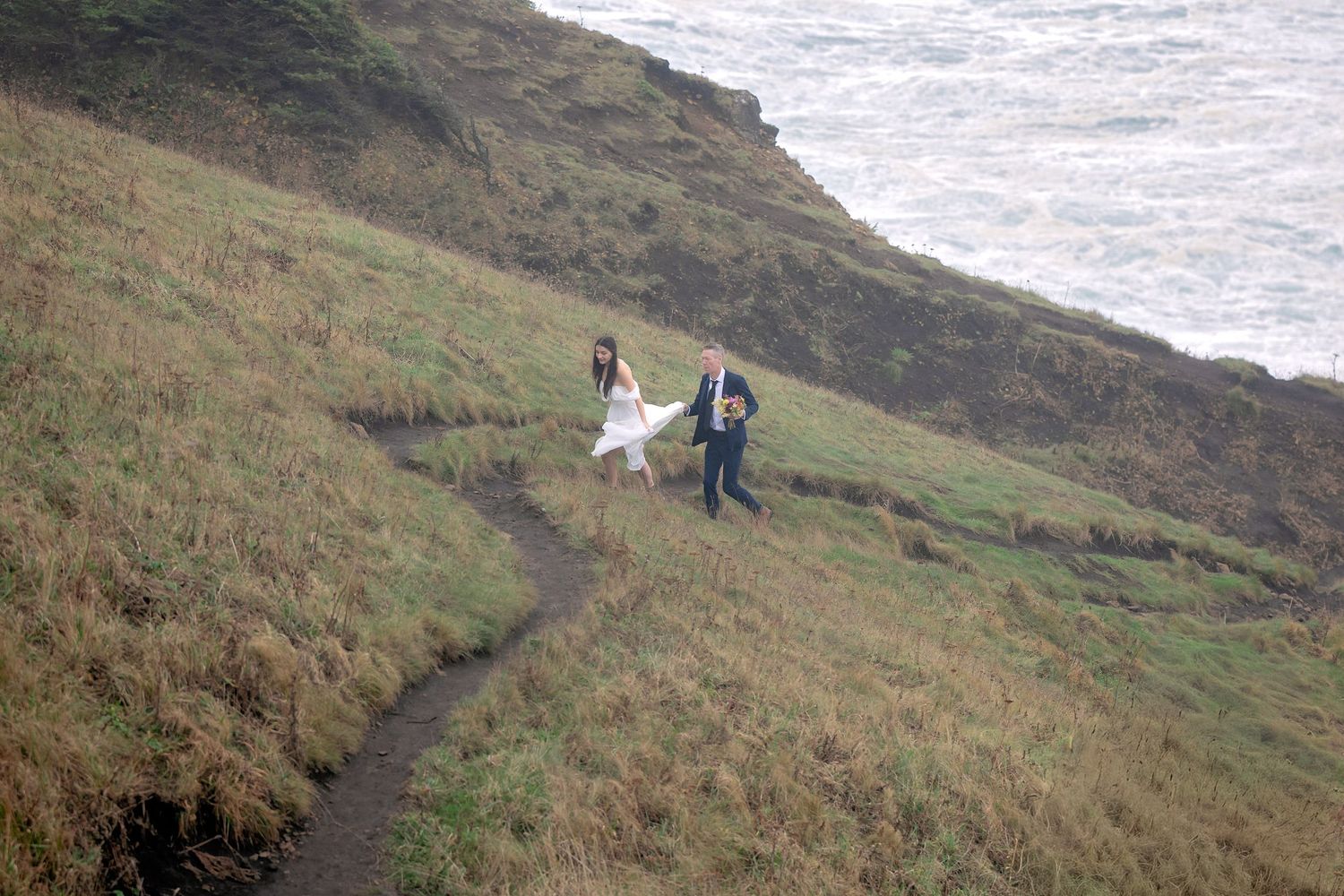 A couple in wedding attire walks along a scenic coastal cliff path overlooking the ocean.