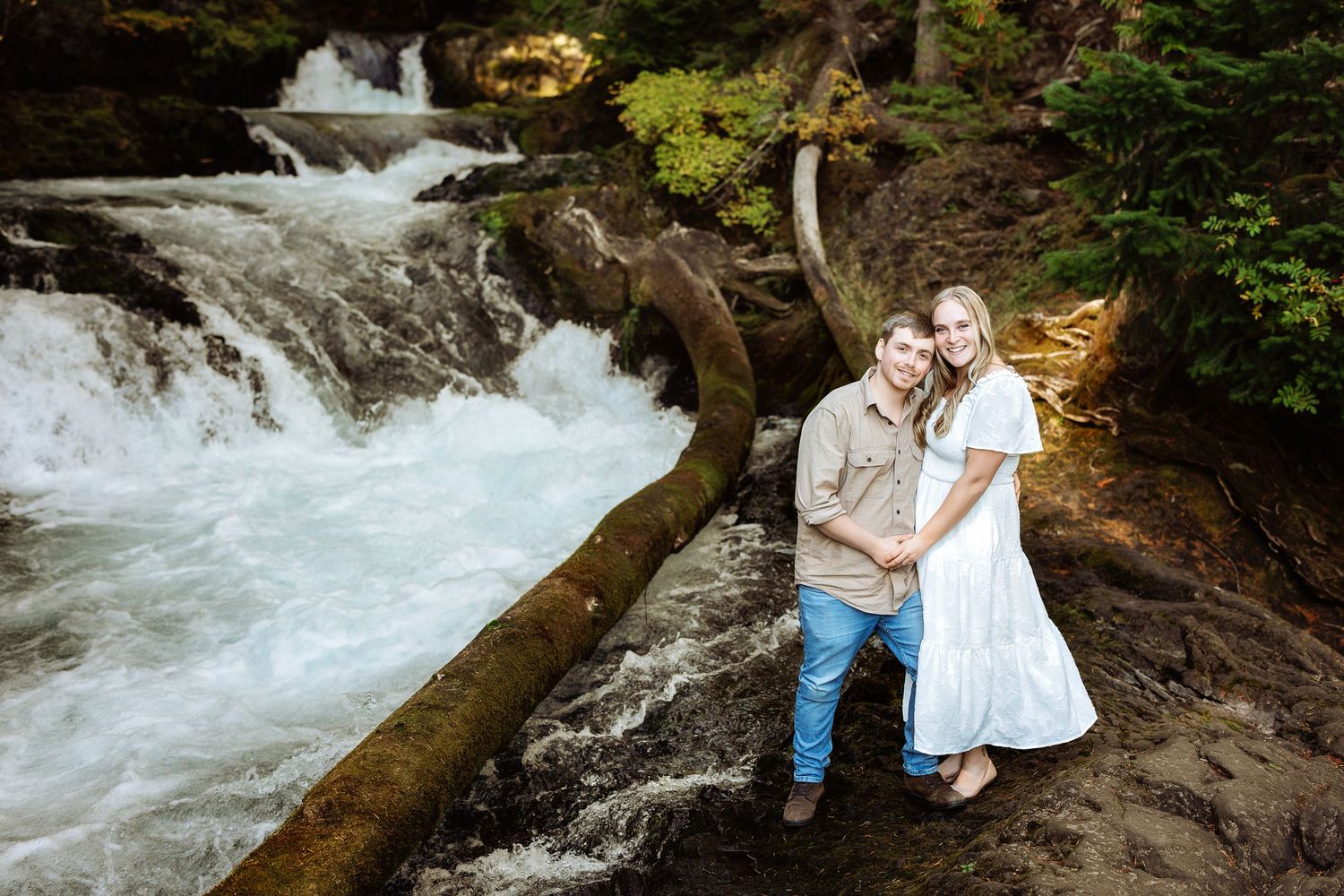 A couple poses together near rushing waterfall cascades in a scenic forest setting.