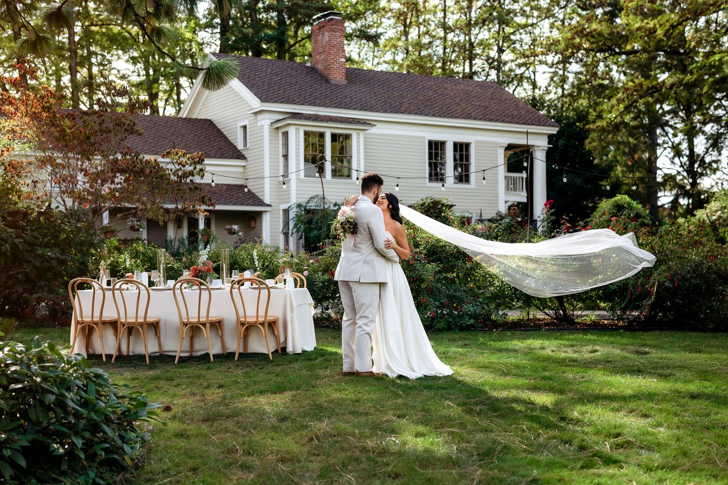 A group of people in formal wedding attire stand in a line on a grassy lawn with a white house in the background.